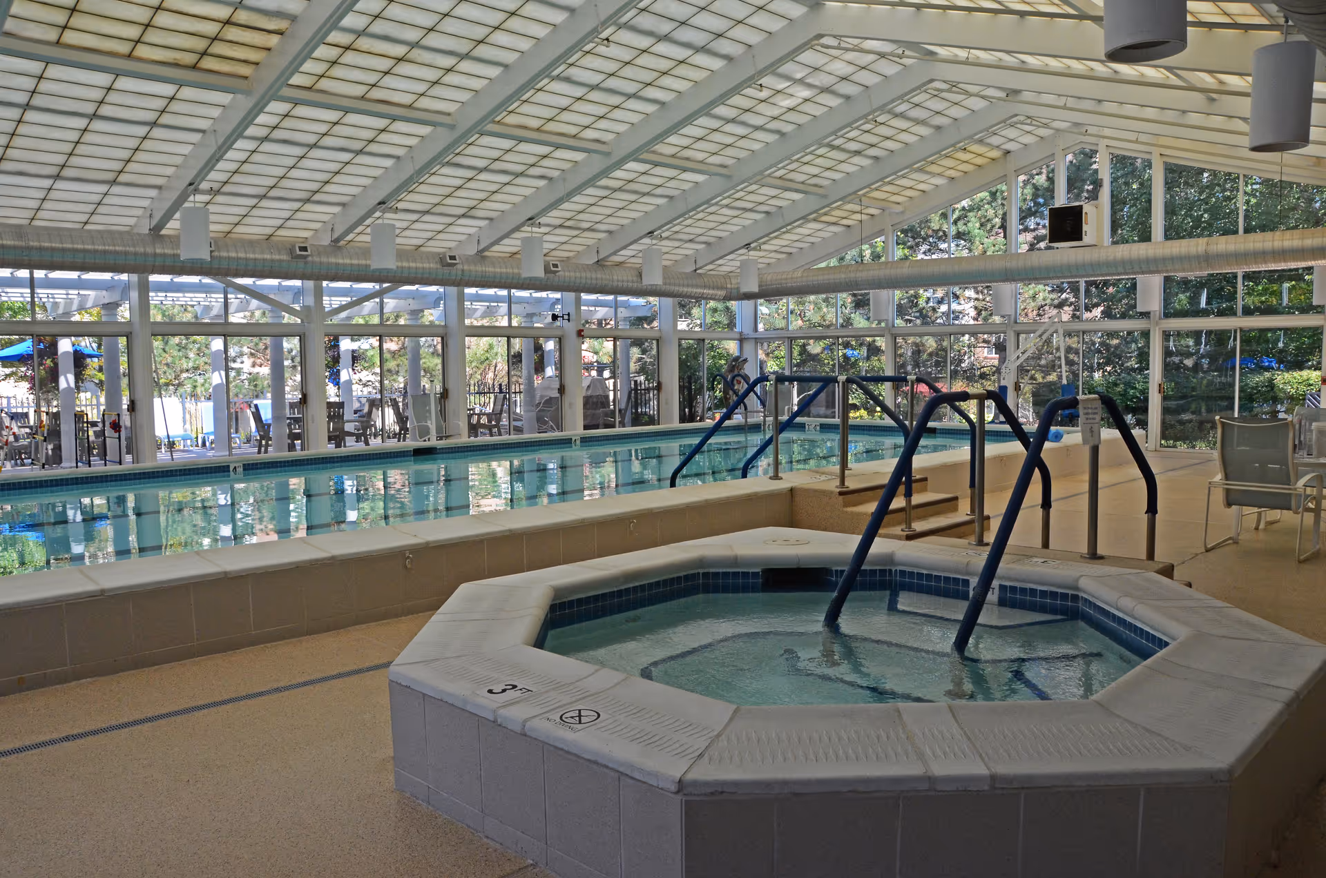 Indoor swimming pool area with a hot tub in the foreground. The pool is surrounded by large windows letting in natural light, with tables and chairs visible outside the pool area.