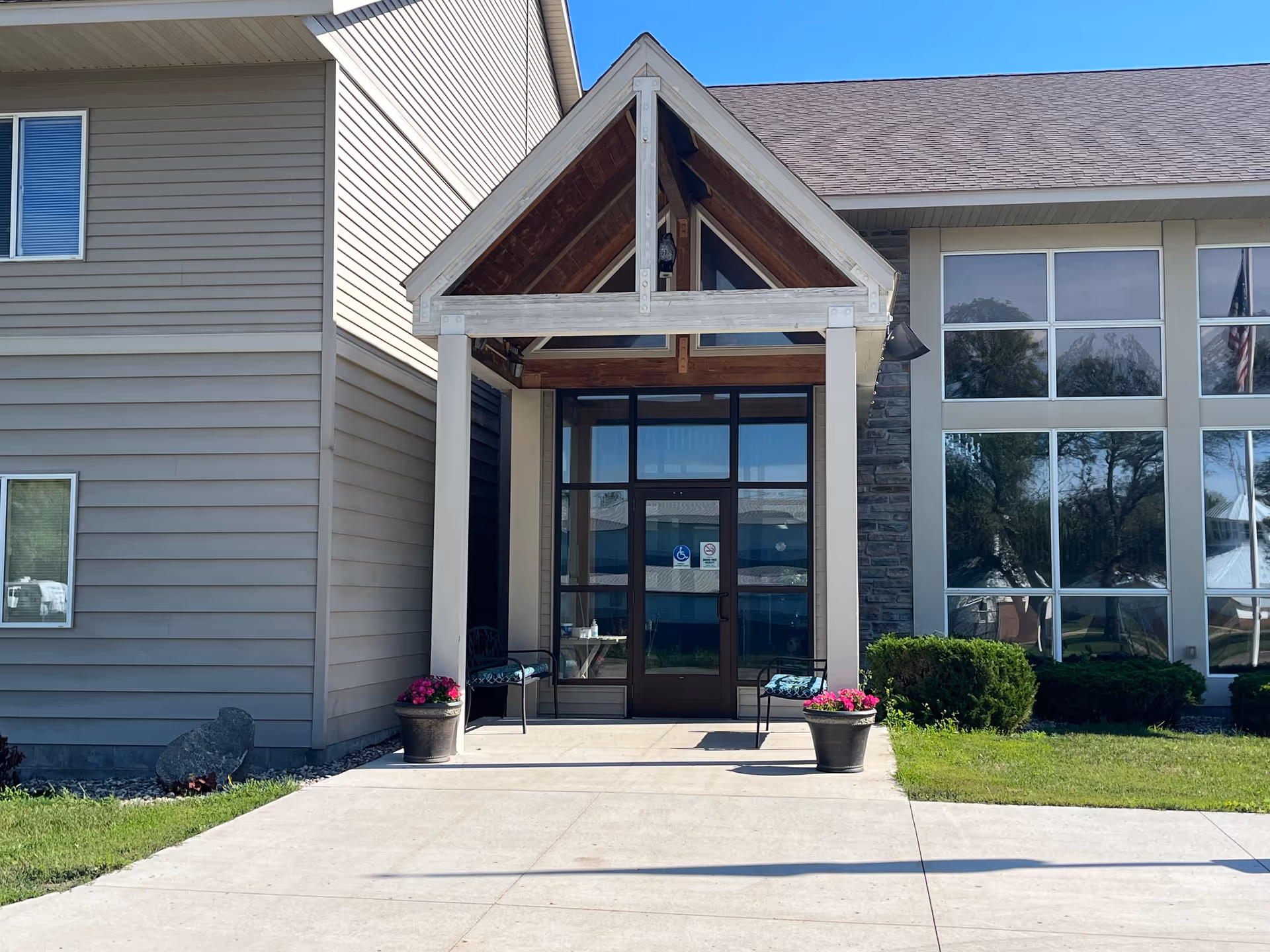 Entrance of a building with a peaked roof overhang supported by two columns. There are two black chairs with cushions and two flower pots with pink flowers on either side of the entrance door. The building has large windows on the right side and beige siding on the left side. The sky is clear and blue.
