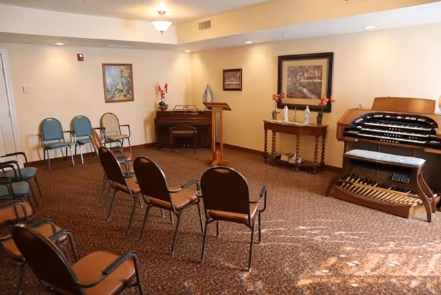 A small meeting room with rows of chairs facing a podium, a piano and an organ under warm lighting.