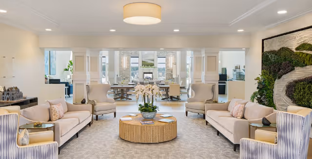 A spacious and well-lit living room area with two beige sofas, four armchairs, and a round wicker coffee table with a flower arrangement in the center. The room features a neutral color palette with light walls and carpet, recessed lighting, and a large decorative wall piece with greenery on the right. In the background, there is a dining area with tables and chairs, and large windows letting in natural light.