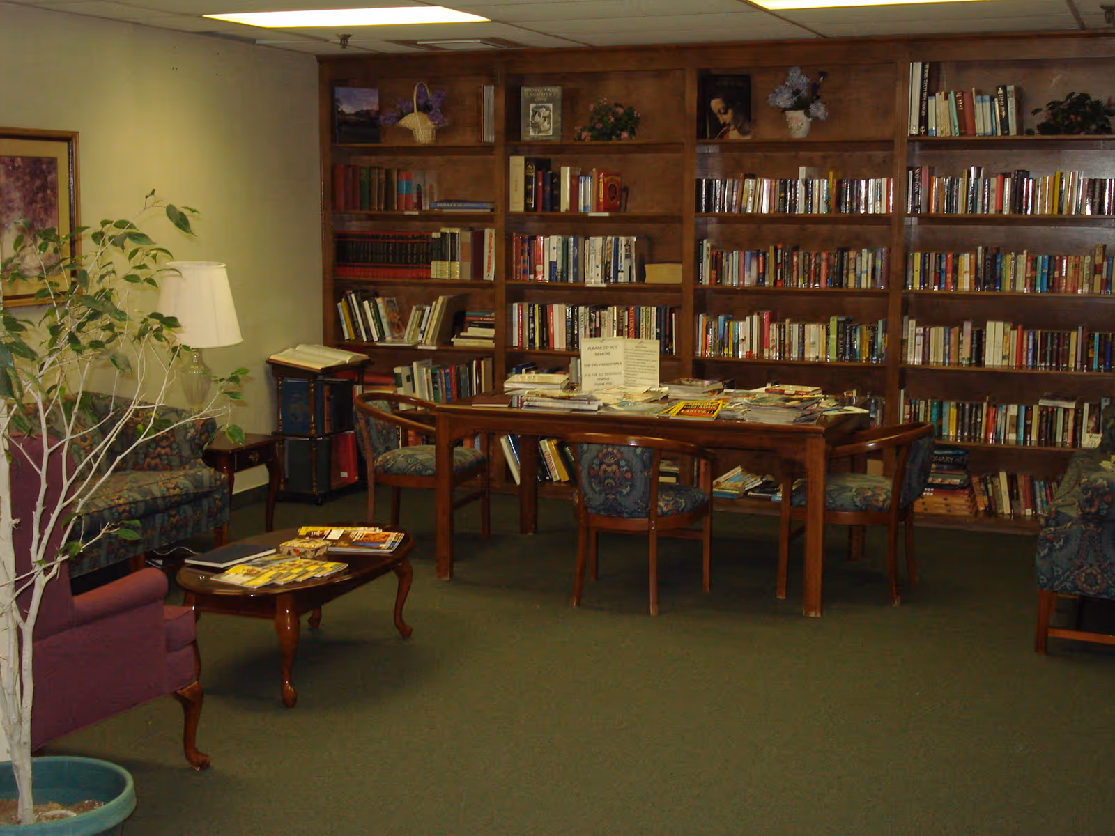 A cozy library room with wooden bookshelves filled with books along the back wall. In front of the shelves is a wooden table covered with magazines and papers, surrounded by four upholstered chairs with floral patterns. To the left, there is a purple armchair, a small wooden coffee table with magazines, a floor lamp, and a potted plant. The room has a green carpet and soft lighting from ceiling fixtures.