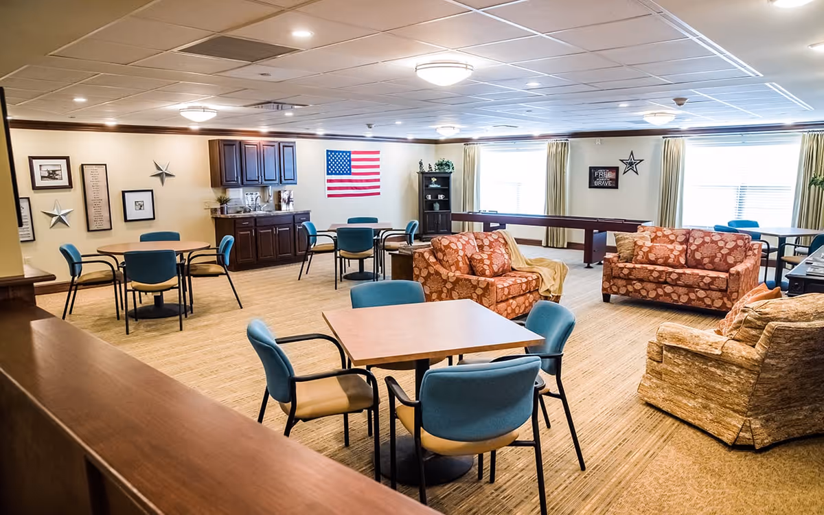A spacious common area in a senior living facility featuring multiple tables with blue chairs, two patterned sofas, an armchair, and a shuffleboard table near large windows with beige curtains. The walls are decorated with framed pictures, stars, and an American flag. The room has a beige carpet and a ceiling with recessed lighting.