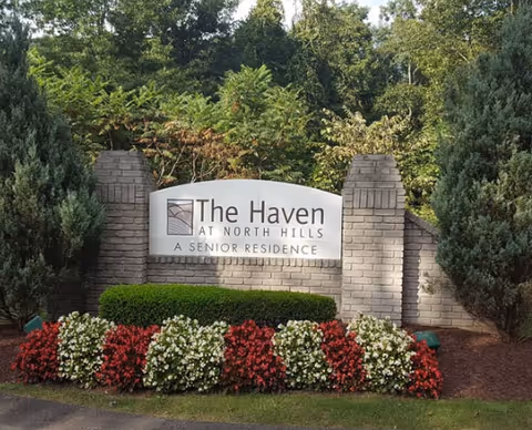 A brick sign at the entrance of The Haven at North Hills Senior Residence surrounded by green bushes and red and white flowers, with trees in the background.