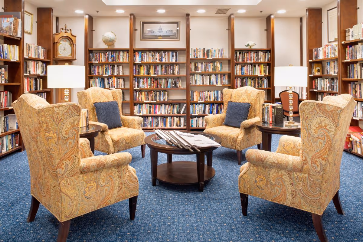 A cozy library room with four patterned armchairs arranged around a round wooden coffee table holding newspapers and magazines. The walls are lined with tall wooden bookshelves filled with books. Two table lamps with white shades are placed on side tables between the chairs. The room has a blue carpet and a framed picture and a globe on the shelves.