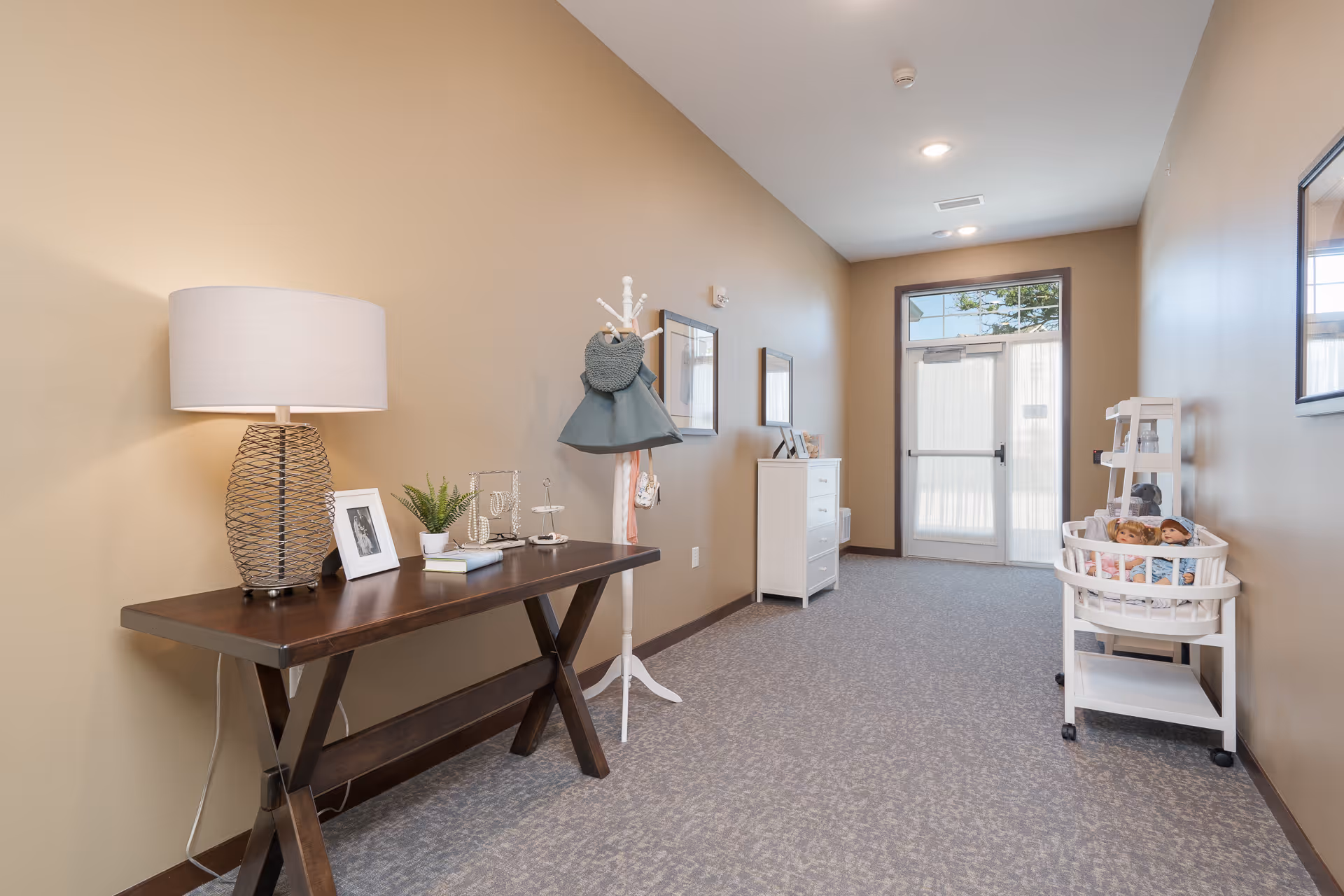 Well-lit interior hallway with a wooden console table and lamp, a coat rack, and a storage cart near a glass exit door.