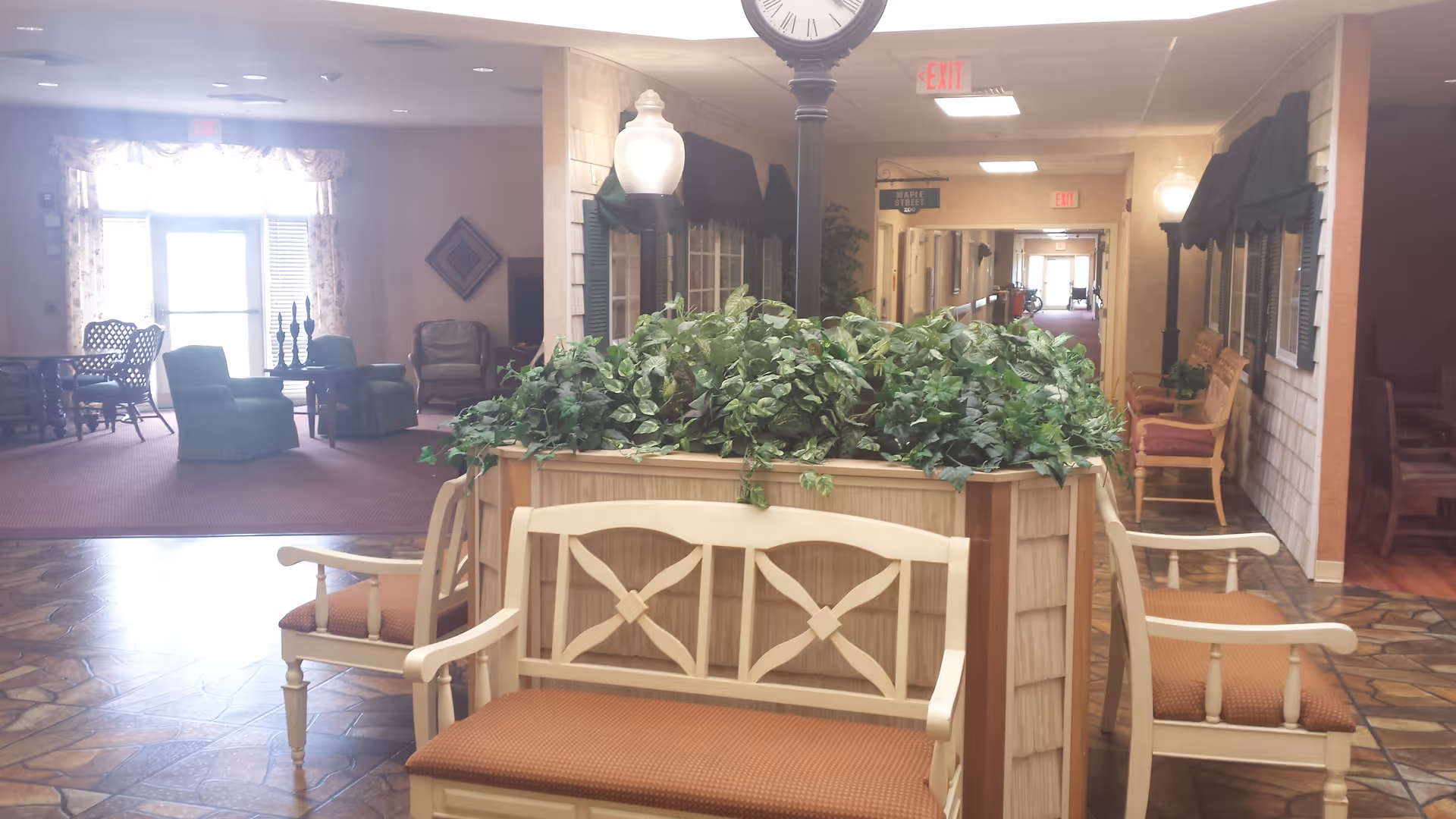 Indoor seating area in a senior living facility with a central planter filled with green plants surrounded by four wooden benches with orange cushions. In the background, there are additional chairs and tables, a hallway with exit signs, and a clock mounted on a post above the planter.