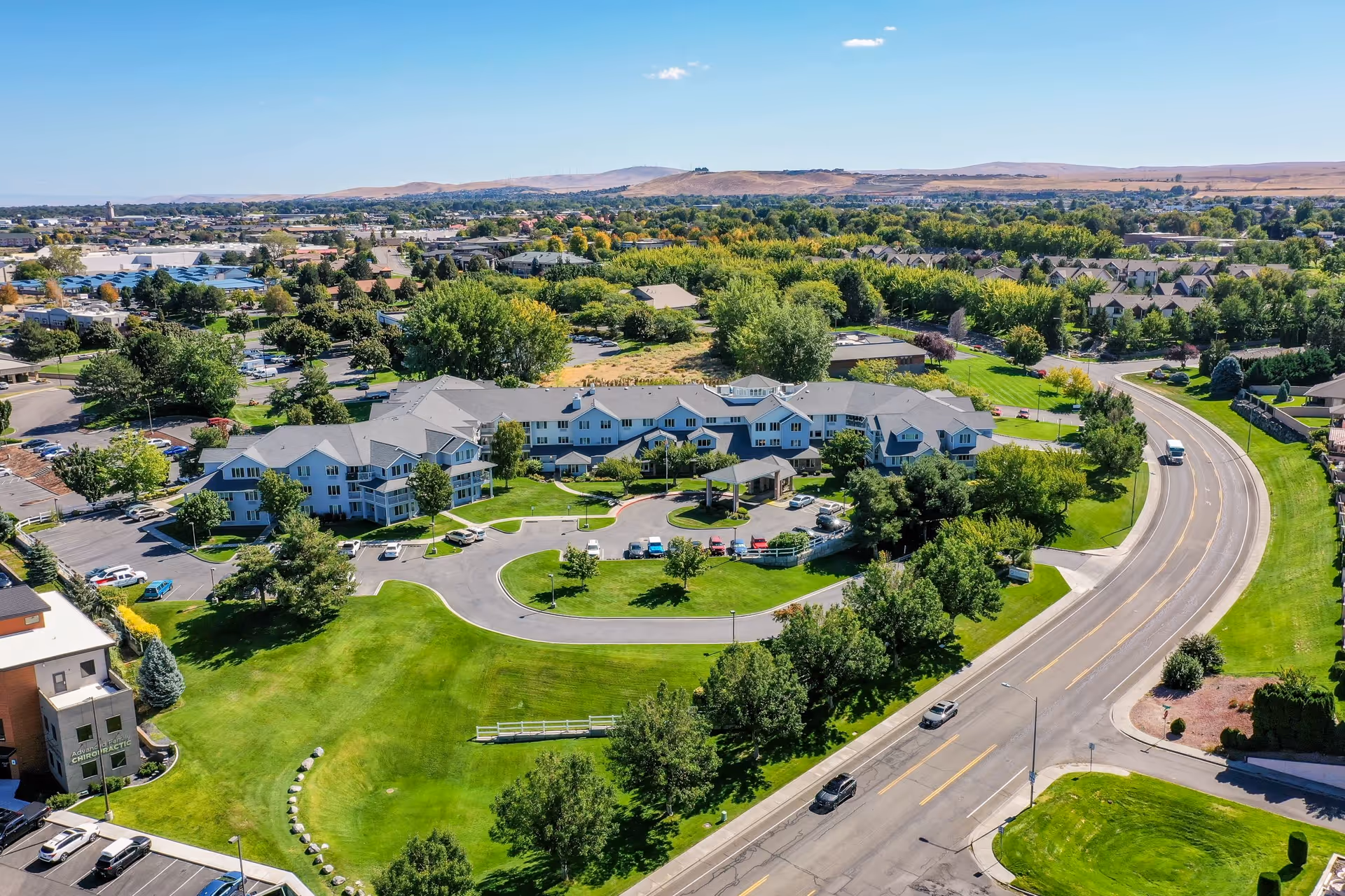 Aerial view of Solstice Senior Living at Kennewick facility surrounded by green lawns, trees, and a curved road with cars. The building is a large, multi-wing structure with a parking area and a covered entrance. The surrounding area includes other buildings, roads, and distant hills under a clear blue sky.