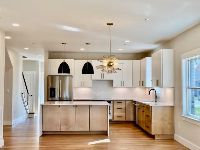 Modern kitchen with light wood lower cabinets and white upper cabinets, stainless steel refrigerator and dishwasher, a kitchen island with two black pendant lights above, and a large window on the right letting in natural light.
