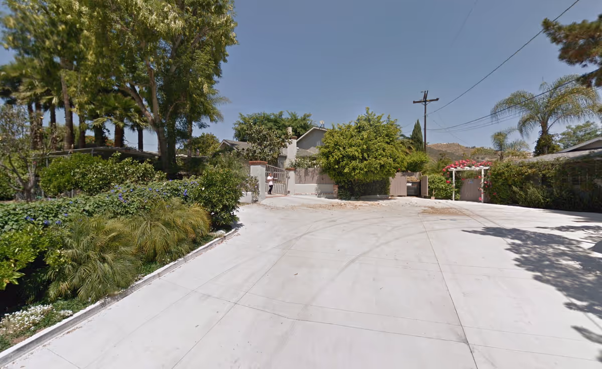 Wide concrete driveway leading to a gated residential entrance flanked by trees and shrubs under a clear sky.
