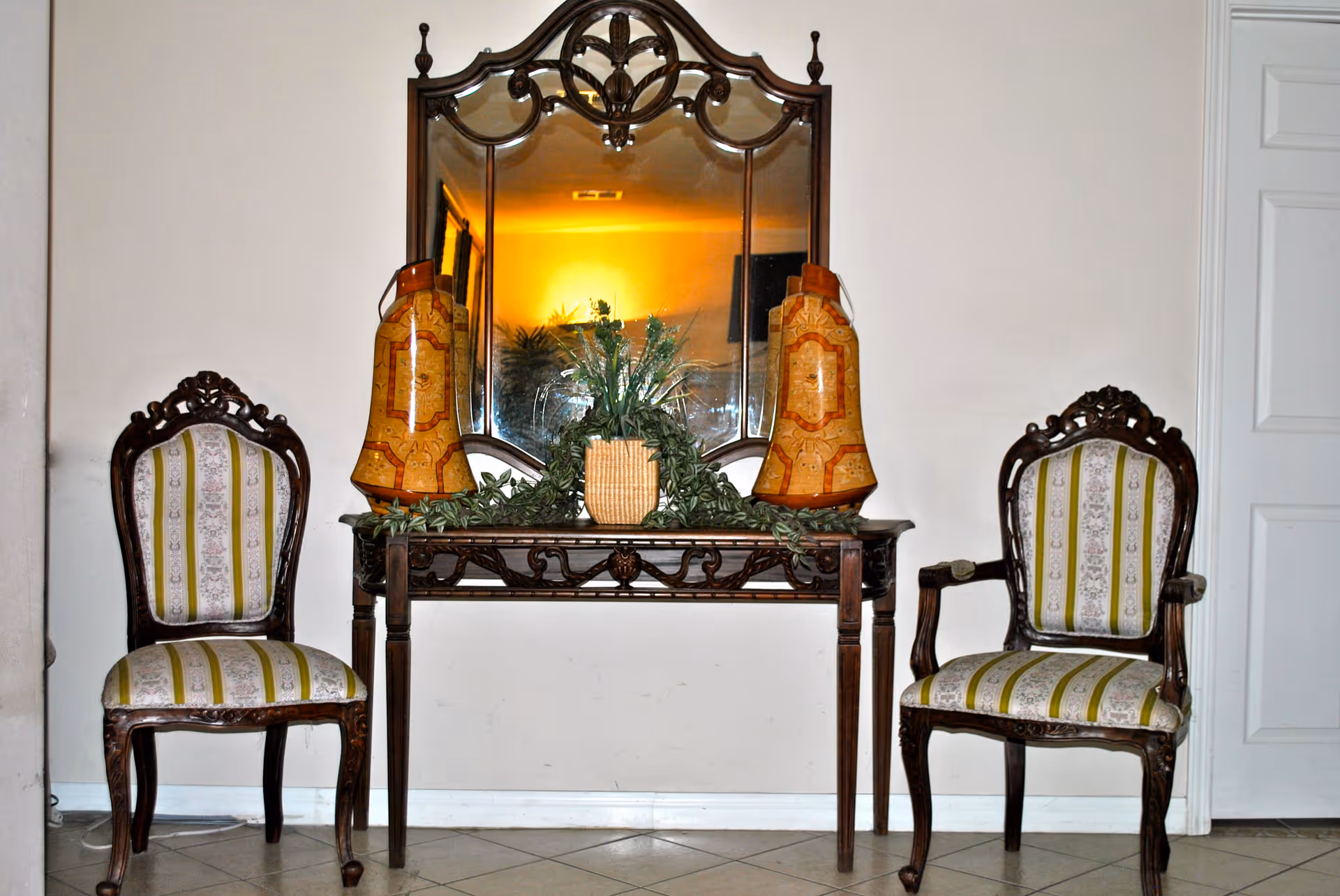An interior setting featuring two ornate wooden chairs with striped upholstery placed on either side of a wooden console table. The table holds a decorative plant in the center and two large decorative vases on each side. Above the table is a large, intricately framed mirror reflecting a warm light and some greenery.