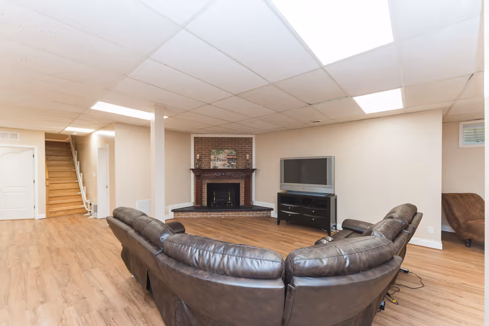 A spacious living room with a curved dark brown leather sectional sofa facing a flat-screen TV on a black TV stand. The room features a brick fireplace with a wooden mantle and a painting above it. The floor is wood laminate, and the ceiling has white tiles with recessed lighting. There is a staircase with a handrail leading upstairs and a white door next to it.
