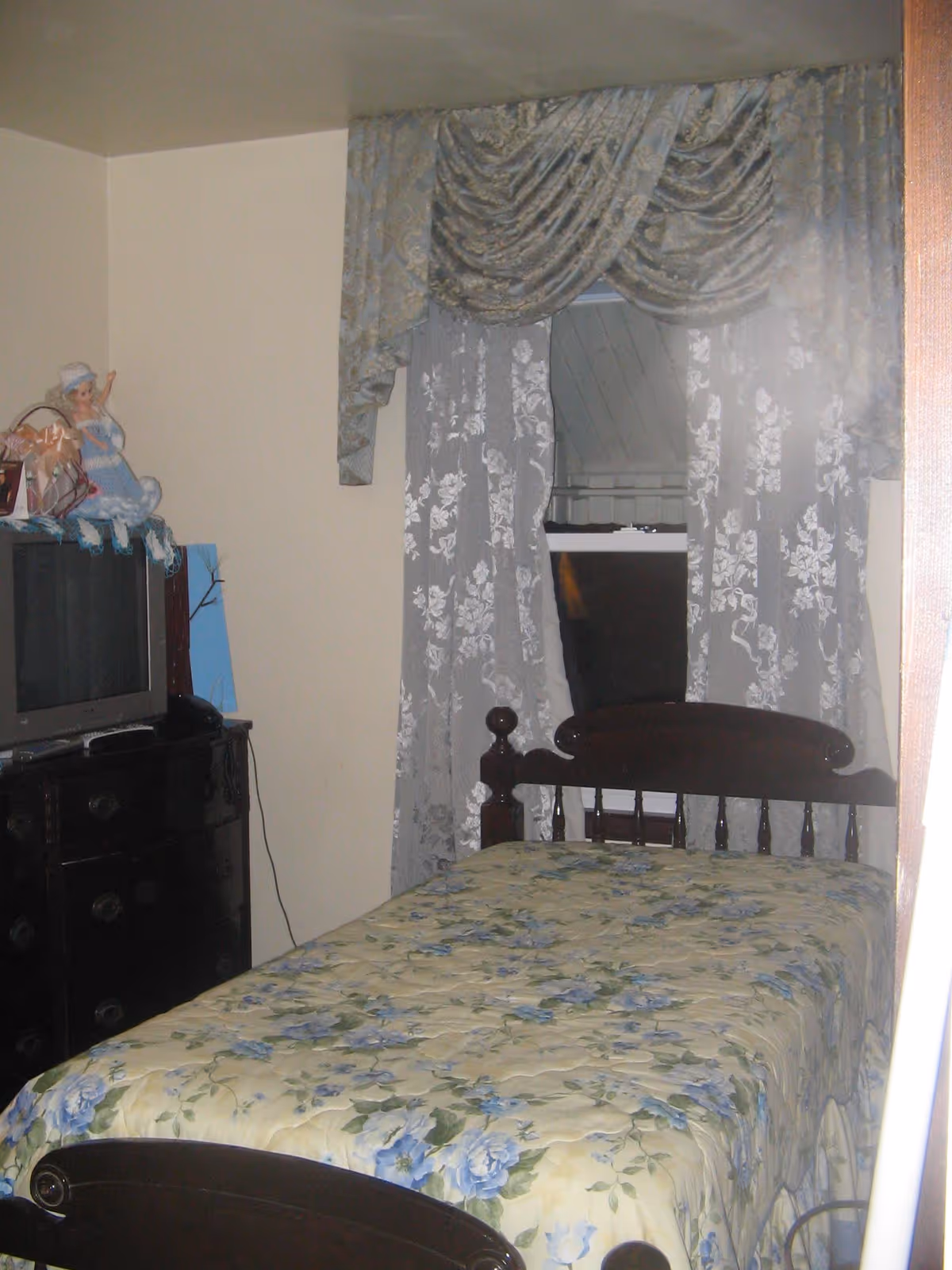 A small bedroom with a single bed covered in a floral bedspread with blue and green flowers. The bed has a dark wooden headboard and footboard. Next to the bed is a dark wooden dresser with a small TV on top. A doll dressed in blue sits on the dresser. The window behind the bed is covered with lace curtains and a decorative valance.