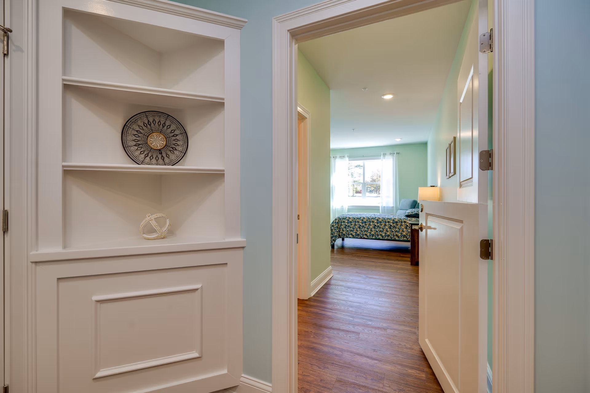 View through an open door into a bedroom with a bed covered in a floral bedspread, a bedside table with a lamp, and a window with sheer curtains letting in natural light. In the foreground, there is a built-in white shelving unit with decorative items on the shelves.