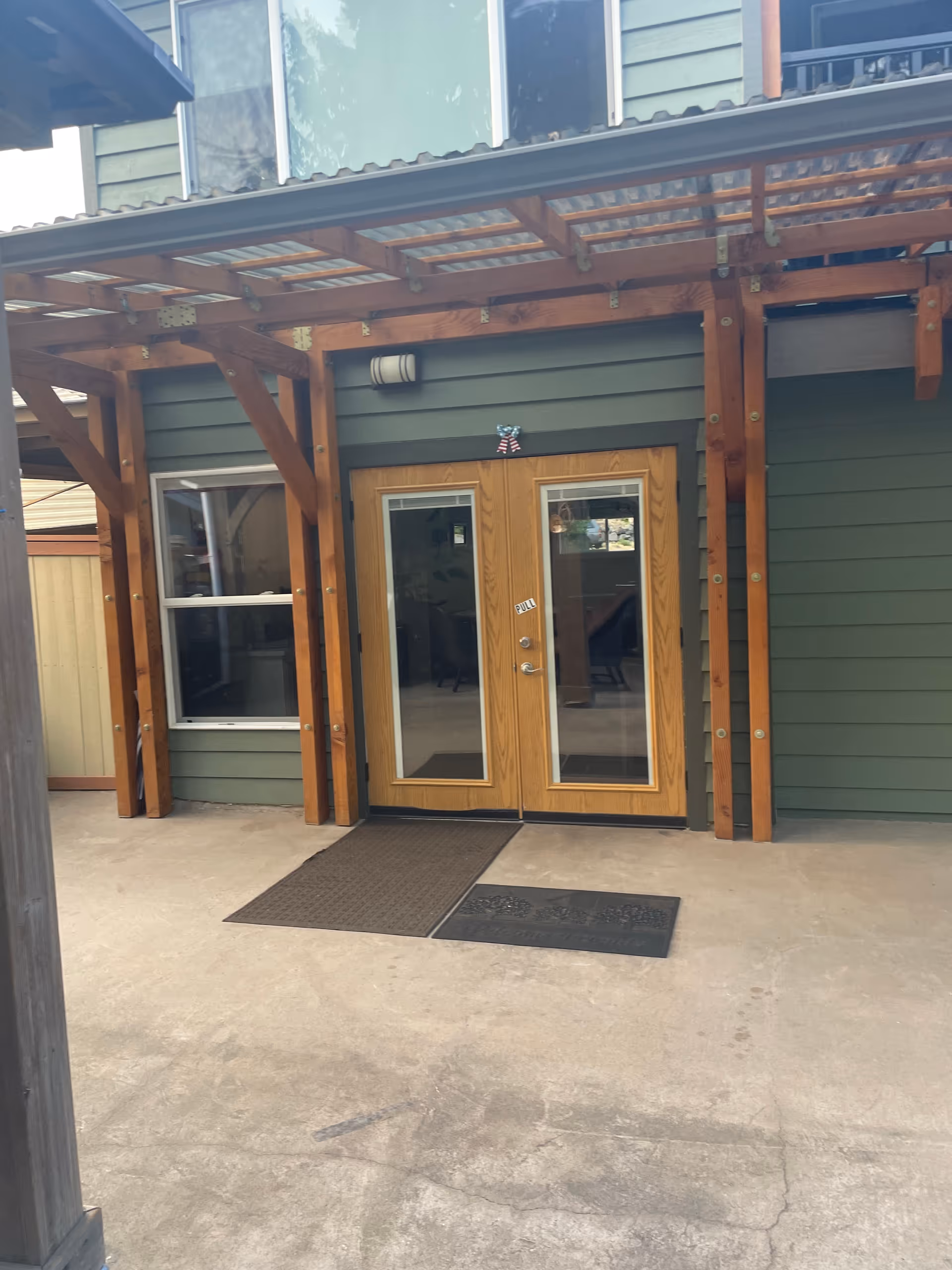 Exterior view of a building entrance with double wooden doors featuring glass panels. The doors are under a wooden pergola structure with a translucent roof. There are two doormats placed on the concrete ground in front of the doors, and a window is visible to the left of the doors.