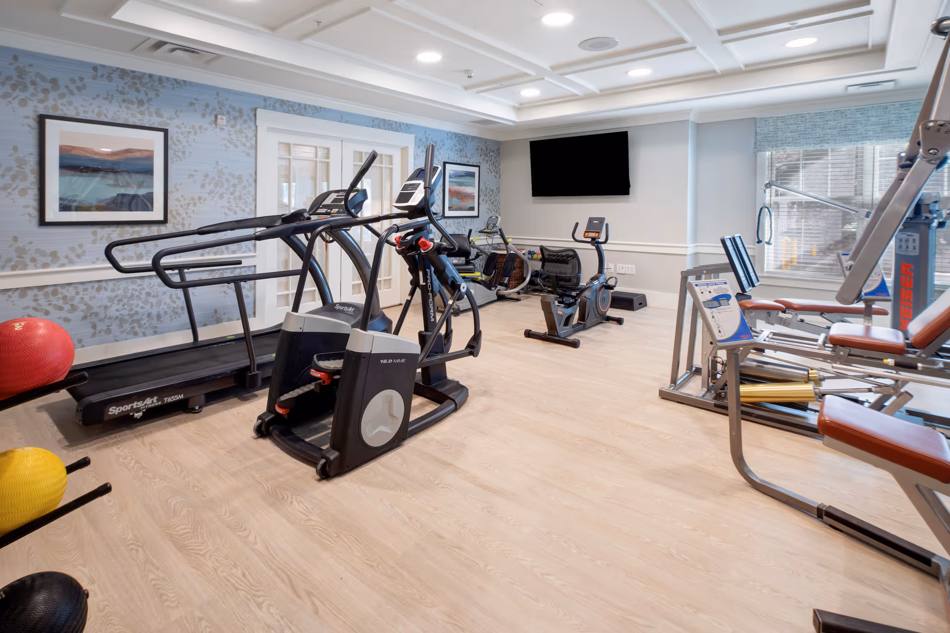 A well-lit fitness room with exercise equipment including a treadmill, elliptical machine, stationary bike, and weight machine. The room has light wood flooring, light blue patterned wallpaper, framed artwork on the walls, a large flat-screen TV mounted on the wall, and windows with blue blinds letting in natural light.