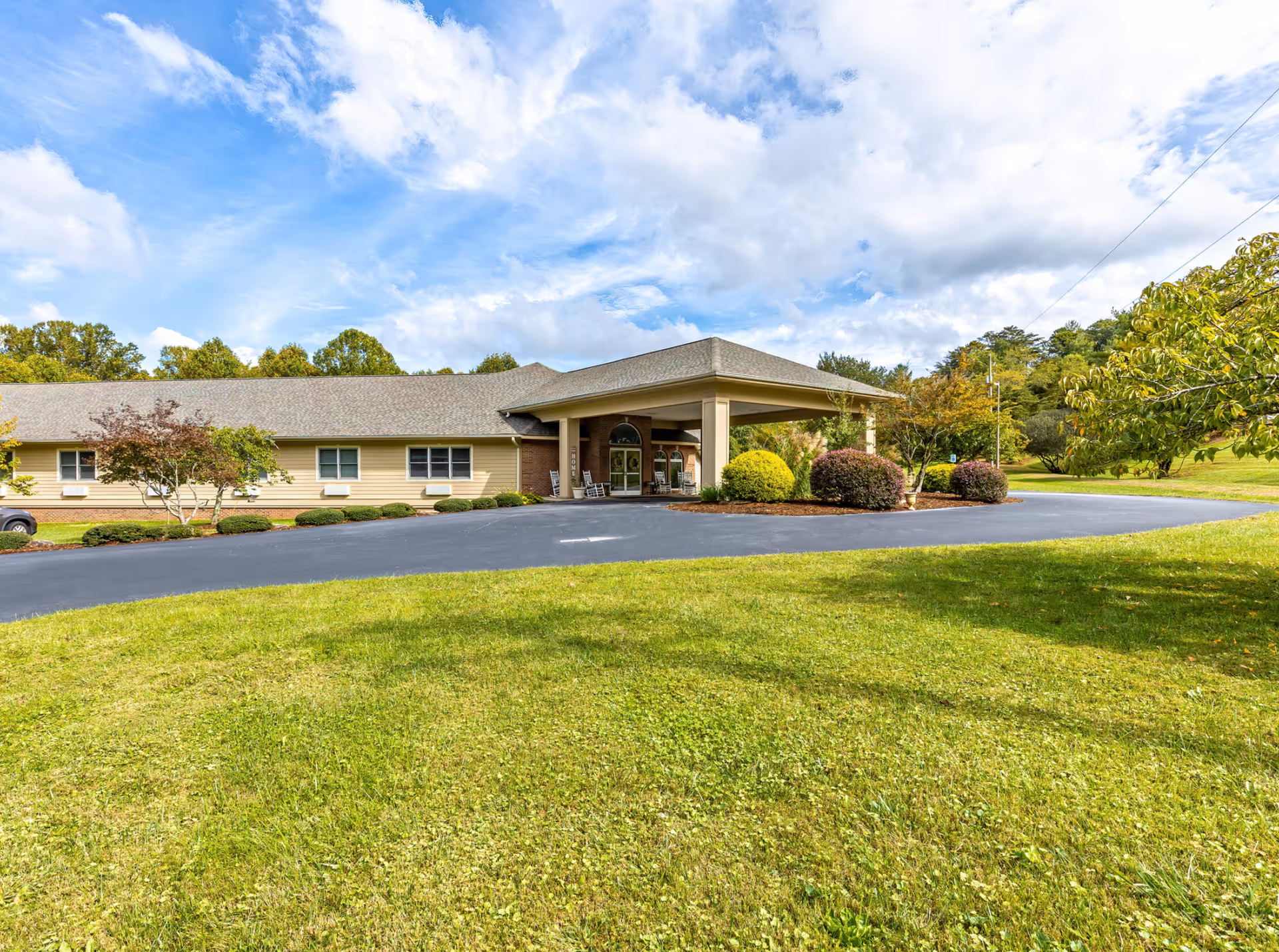 Exterior view of Cherry Springs Village facility showing a single-story building with a covered entrance, surrounded by green grass, bushes, and trees under a partly cloudy blue sky.