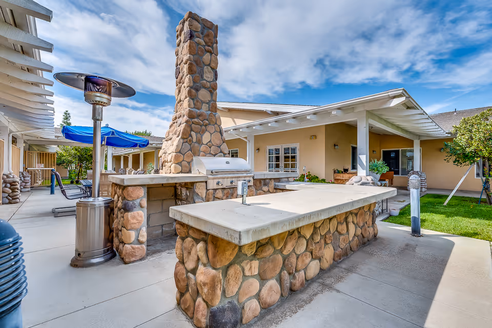Outdoor courtyard featuring a stone-built barbecue island and chimney with patio seating, umbrellas, and covered walkways.