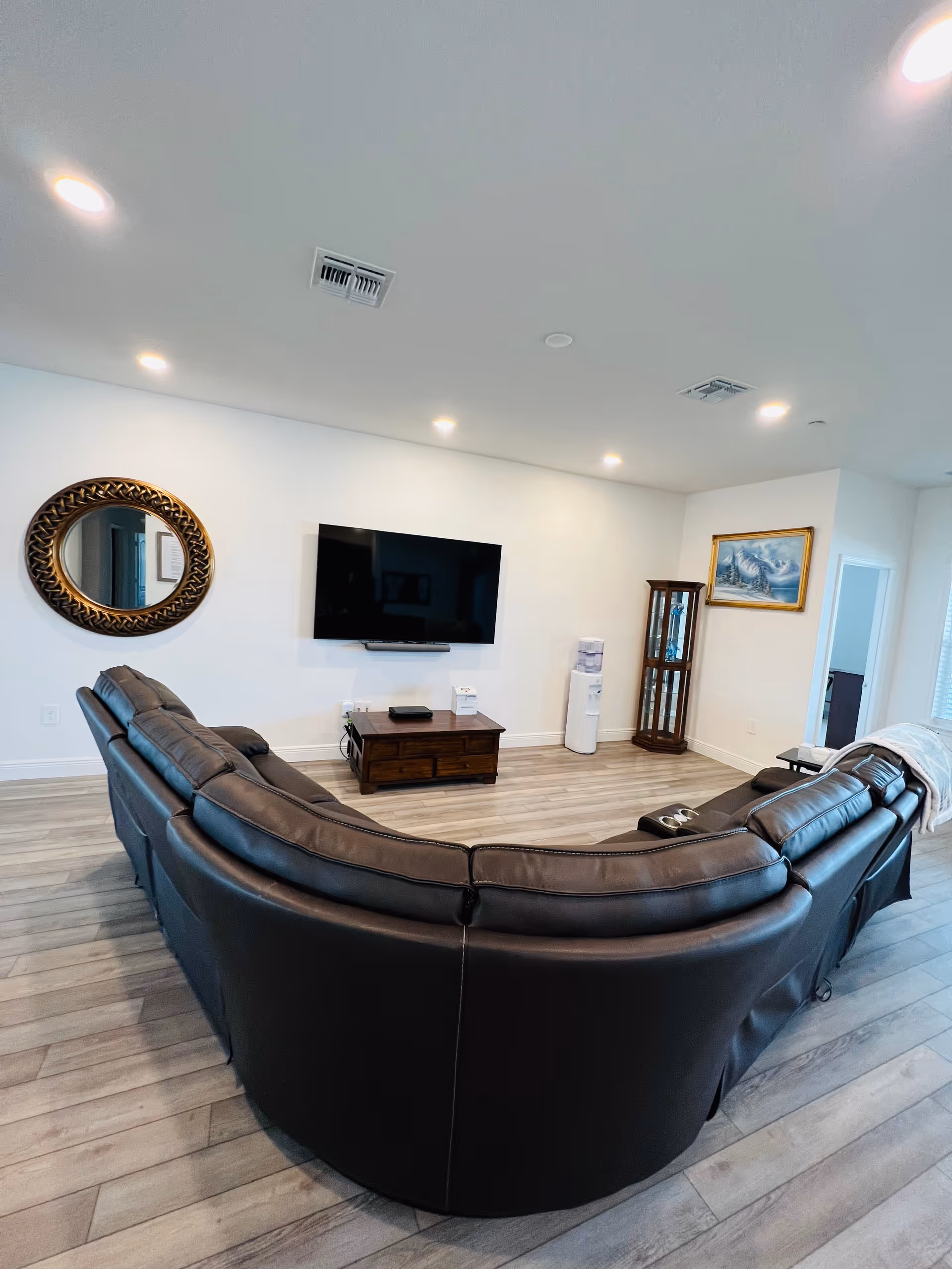 A spacious living room with a large curved black leather sectional sofa facing a wall-mounted flat-screen TV. The room has light-colored wooden flooring, a round decorative mirror on the left wall, a wooden cabinet under the TV, a water dispenser, a glass display cabinet, and a framed painting on the right wall. The ceiling has recessed lighting.