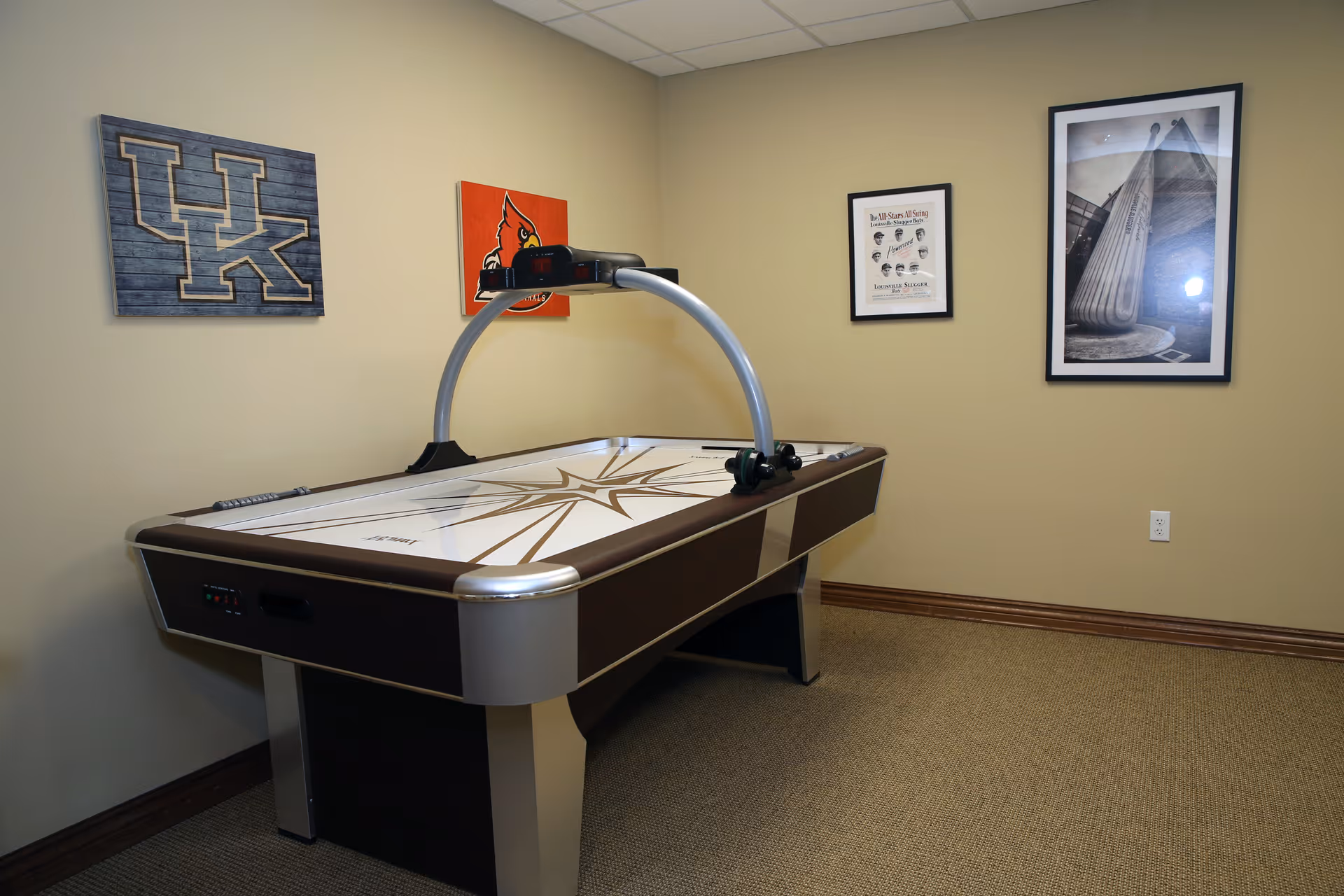 A room with an air hockey table in the center. The walls are decorated with framed pictures and sports-themed artwork, including a UK logo and a cardinal bird. The room has beige walls and carpeted flooring.