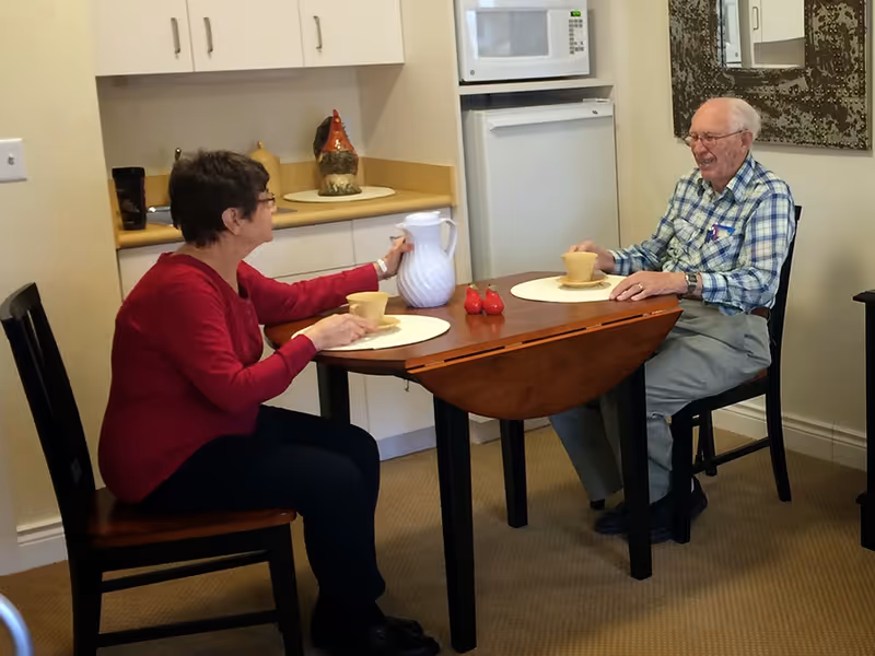 Two older adults sit at a small dining table in a kitchenette area, sharing drinks and smiling.