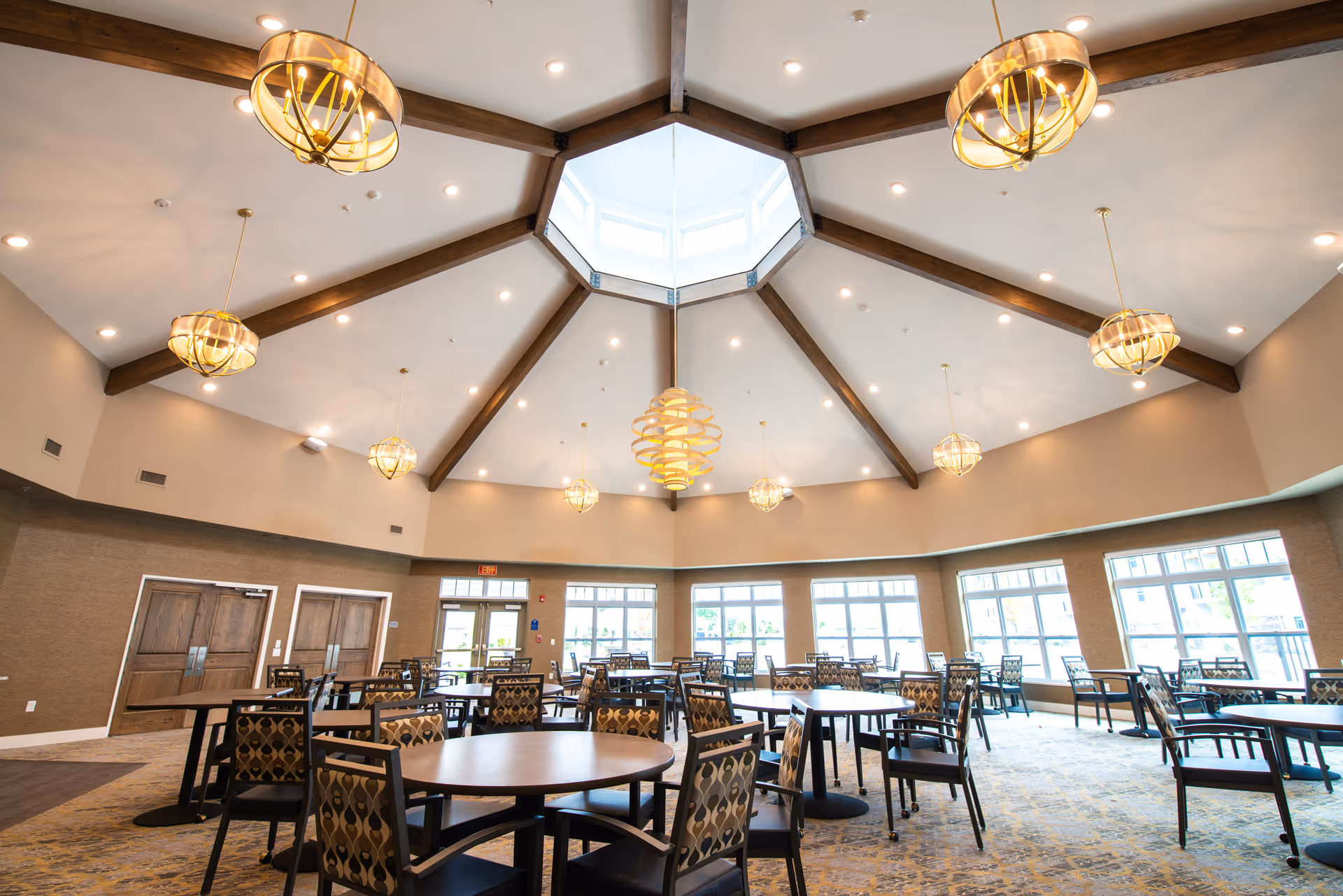 Spacious dining room with multiple round and rectangular tables surrounded by patterned chairs. The room features a high ceiling with wooden beams radiating from a central skylight, and several modern pendant lights hanging down. Large windows line the walls, allowing natural light to fill the space.