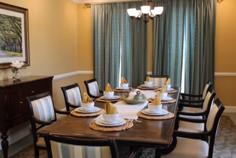 Dining room with a long wooden table set for eight, upholstered chairs, chandelier, sideboard and curtains.