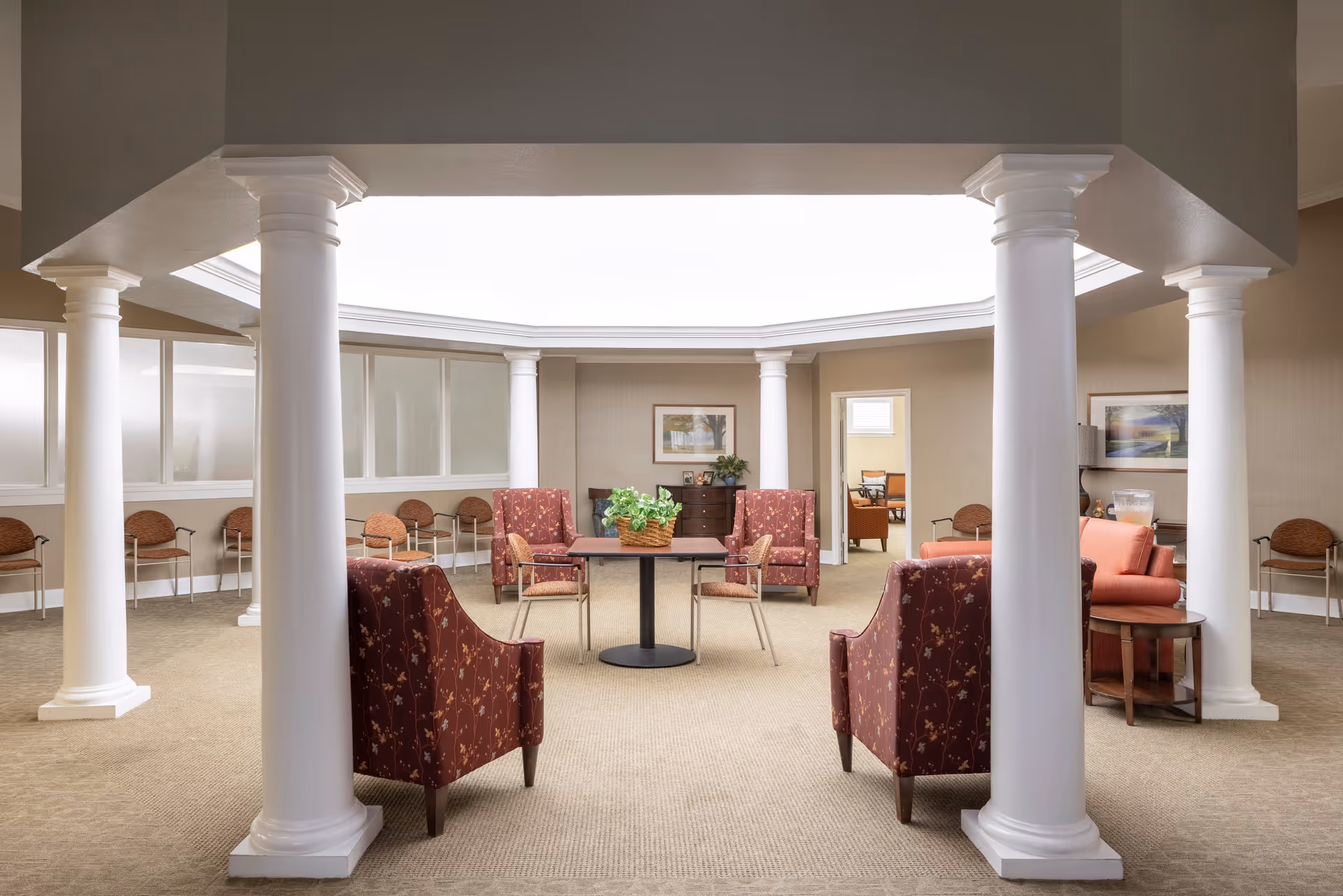 A spacious, well-lit common area with beige walls and carpet, featuring white decorative columns and a large skylight. The room contains several upholstered chairs with floral patterns, a small table with a plant centerpiece, and additional seating along the walls. There are framed pictures on the walls and an open doorway leading to another room.