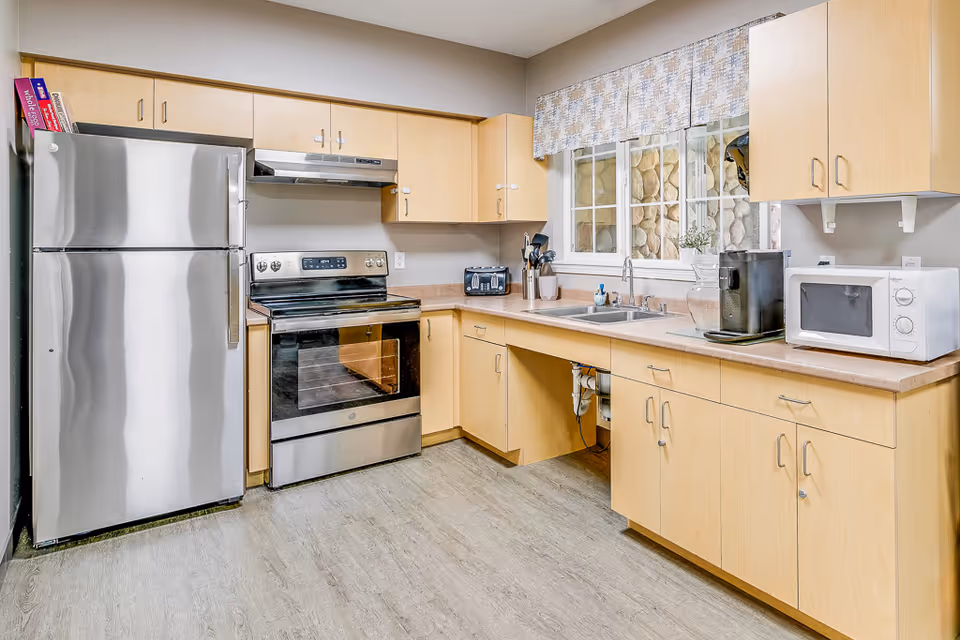 A clean and modern kitchen with light wood cabinets, a stainless steel refrigerator and oven, a double sink under a window with a patterned valance, a microwave, a coffee maker, and various kitchen utensils on the countertop.