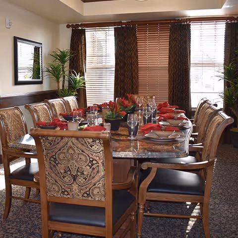 A dining room with a large rectangular table set for a meal. The table has a marble-like surface and is surrounded by wooden chairs with patterned upholstery and black seat cushions. Each place setting includes a plate, bowl, glassware, and red cloth napkins. There are decorative plants and flowers on the table and in the corners of the room. The room has large windows with wooden blinds and dark patterned curtains, and a mirror hangs on one wall.