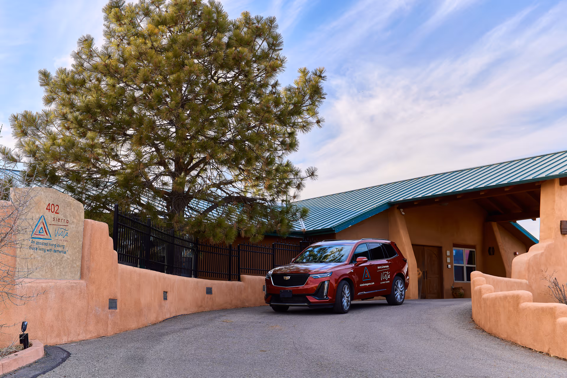 Exterior view of Sierra Vista at Vista Living Care facility showing a driveway with a red SUV parked near the entrance. The building has a tan stucco exterior with a green metal roof, and there is a large tree next to a stone sign displaying the facility name and address 402 Sierra Vista.