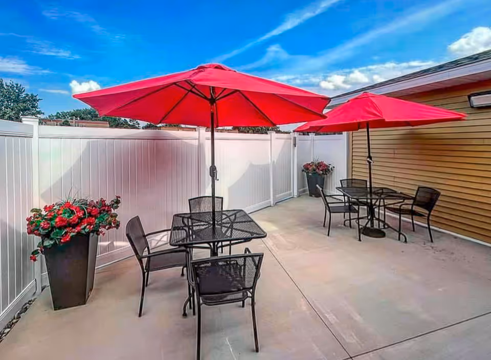 Outdoor patio area with two black metal tables, each with four matching chairs and a large red umbrella. The patio is enclosed by a white fence and the side of a building with tan siding. There are two large planters with red flowers near the fence and building.