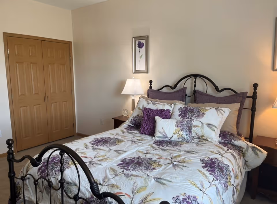 A bedroom with a black metal bed frame, a bed covered with a floral-patterned comforter in shades of purple and green, and multiple pillows. There are two wooden nightstands on either side of the bed, each with a lamp. A framed picture of a purple flower hangs on the wall above the bed. A wooden double-door closet is visible on the left side of the room.