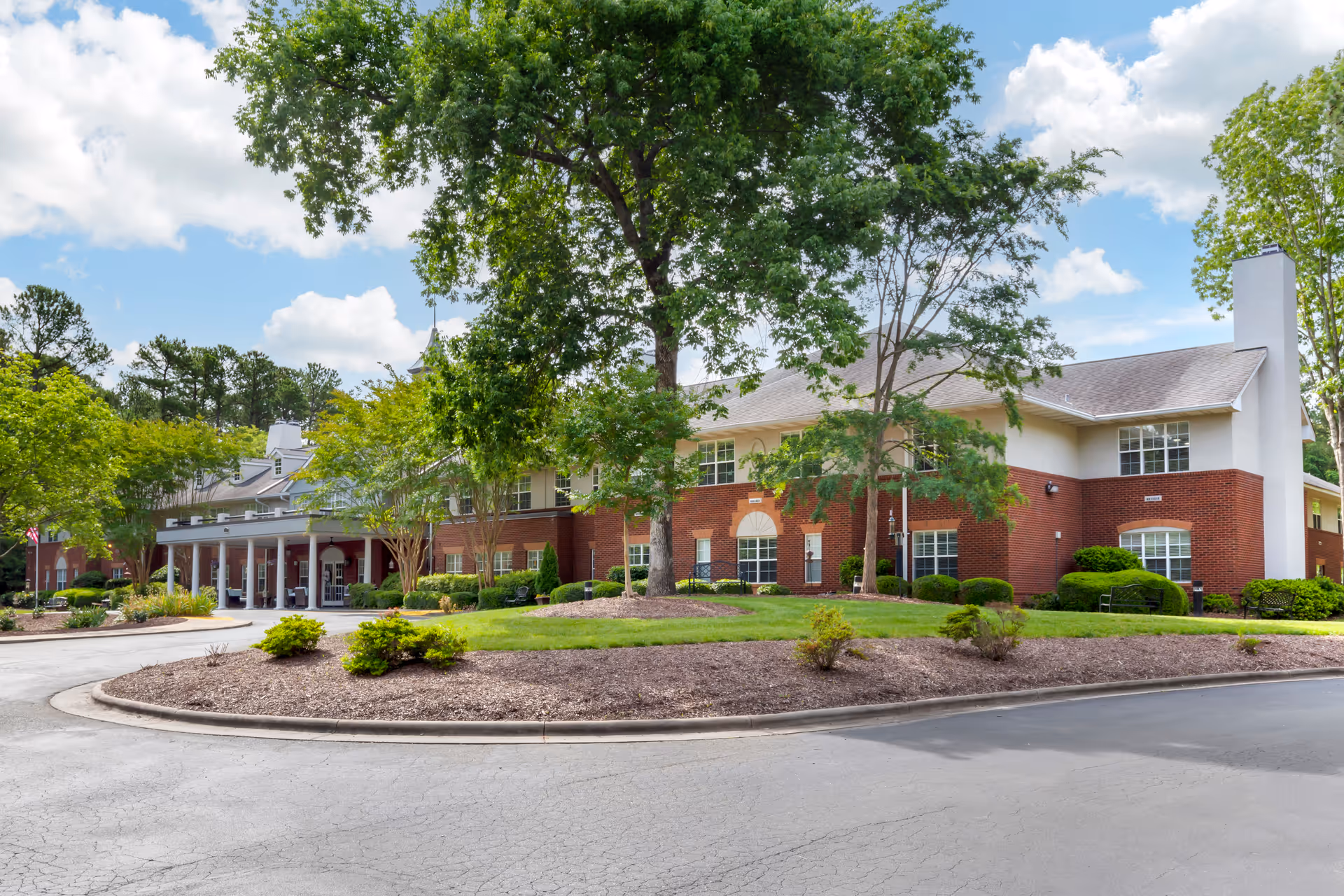 Exterior view of a two-story senior living facility building with red brick and white siding, surrounded by green trees and landscaping under a partly cloudy sky.