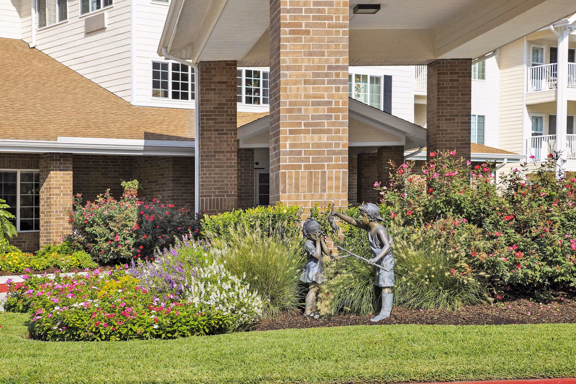 Front entrance of a senior living facility with brick columns, landscaped flower beds, and a bronze statue of two children playing.