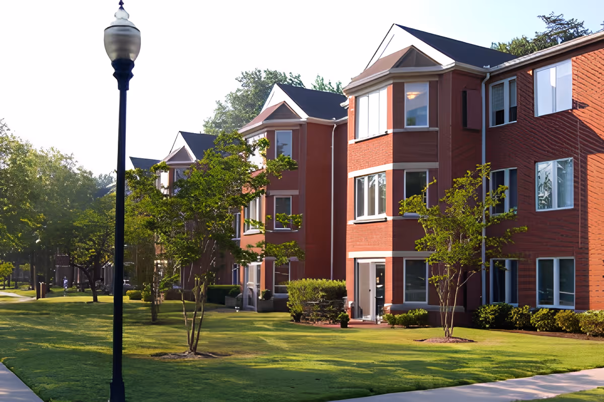 Front exterior of a red-brick multi-story senior living building with a manicured lawn, trees, and a lamppost.