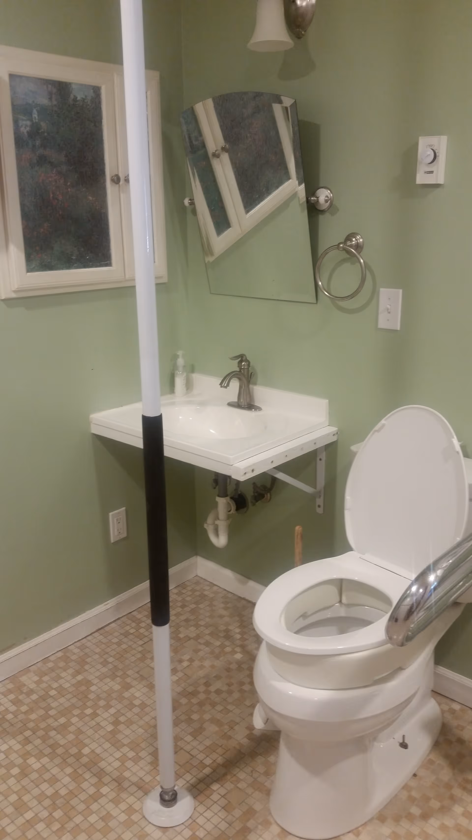 A bathroom with a white toilet equipped with a support rail and a vertical grab bar. There is a white wall-mounted sink with a faucet, a tilted mirror above the sink, a towel ring, and a light switch on the green-painted wall. The floor has small square tiles in beige tones.