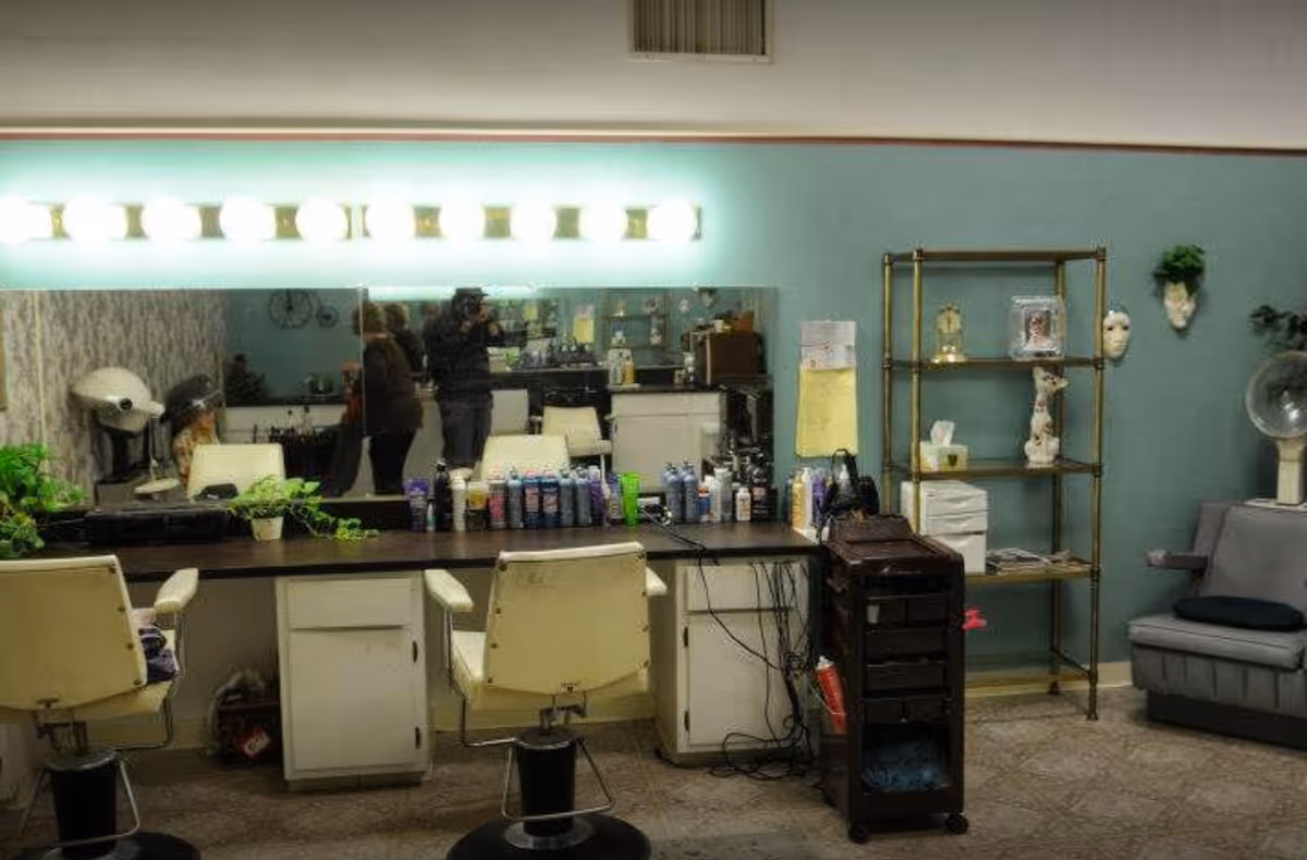 Interior of a hair salon area with two white salon chairs in front of a long counter filled with hair care products. A large mirror with bright lights above it reflects the room, showing a person taking a photo. To the right, there is a metal shelving unit with decorative items and a gray salon hair dryer chair.