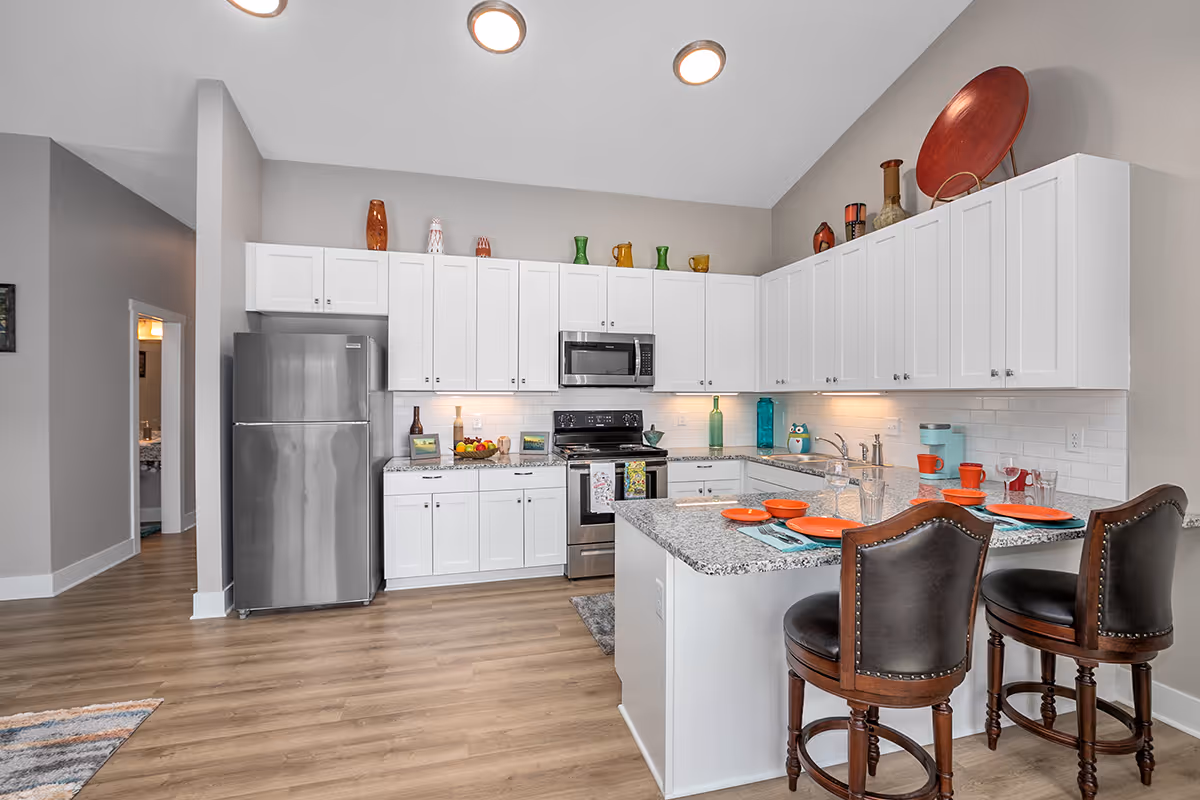 Modern kitchen with white cabinets, granite countertops, stainless steel refrigerator, stove, and microwave. The kitchen island has two dark wooden bar stools with leather seats and is set with orange plates, glasses, and turquoise napkins. Decorative vases and pitchers are displayed on top of the cabinets. The floor is light wood, and the walls are painted light gray.