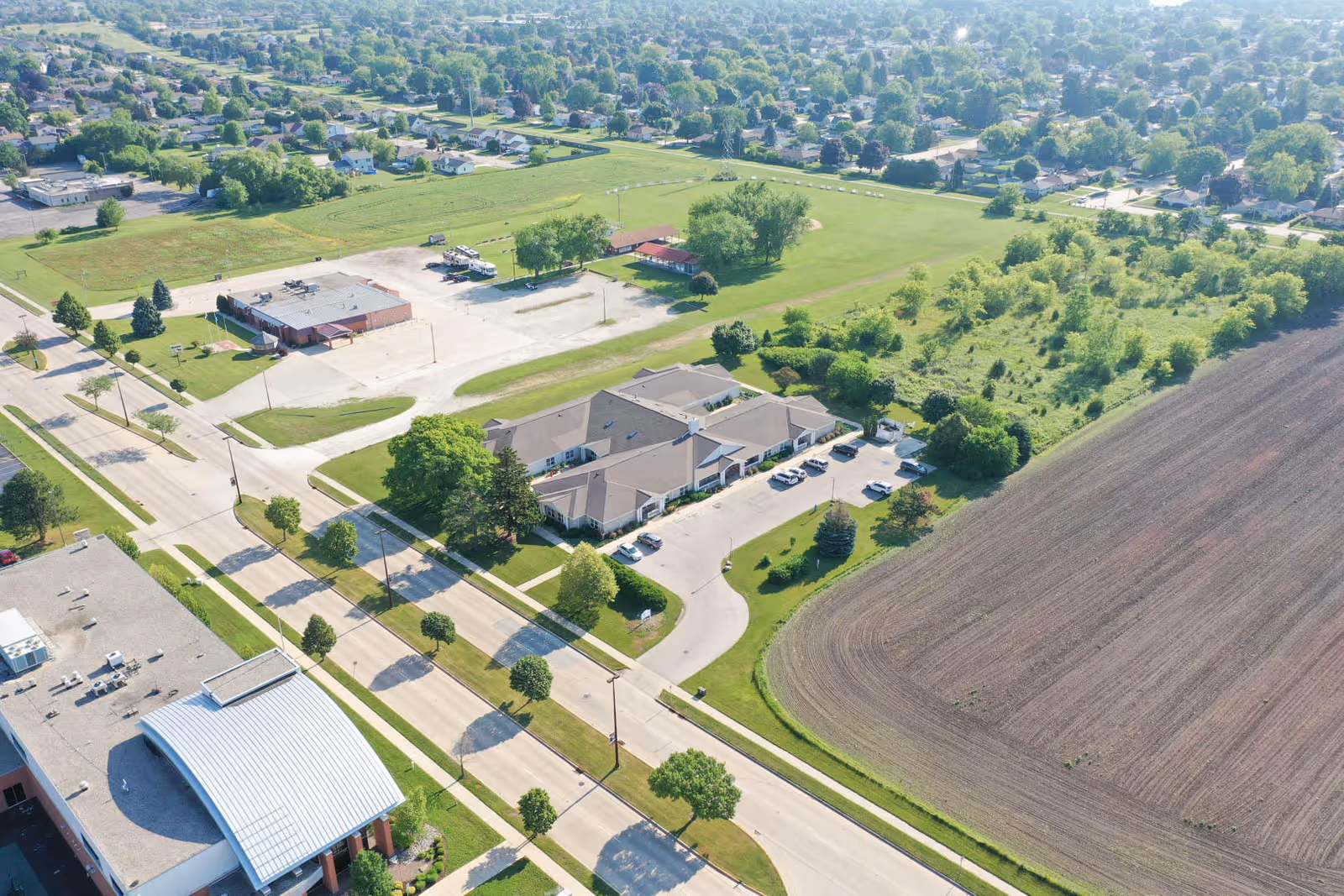 Aerial view of Kenosha Senior Living facility surrounded by green lawns, trees, a large plowed field, and nearby residential neighborhood. The building has a parking lot with several cars and is located near a main road with sidewalks and streetlights.