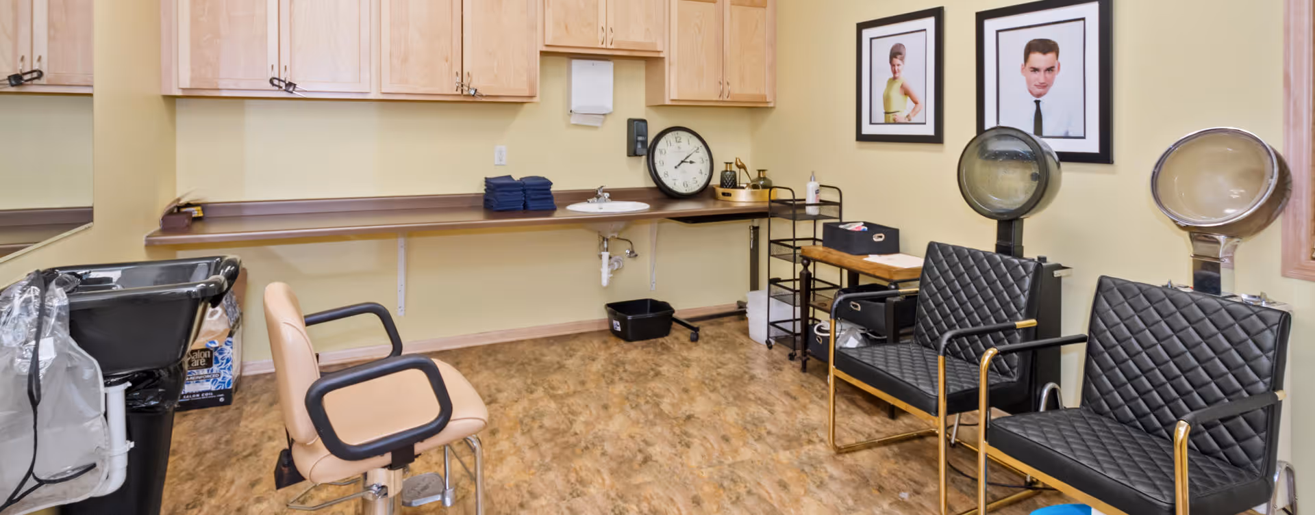 Interior of a salon room with two black salon chairs with hair dryers, a beige salon chair, a black sink for washing hair, wooden cabinets, a countertop with folded towels, a clock, and framed portraits on the wall.