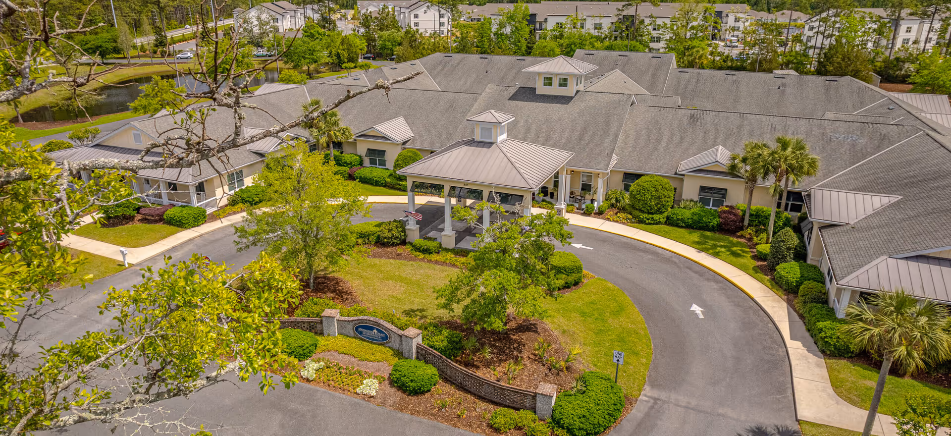 Aerial view of a senior living facility's main entrance with a covered porte-cochere, circular driveway, and landscaped grounds around connected buildings.