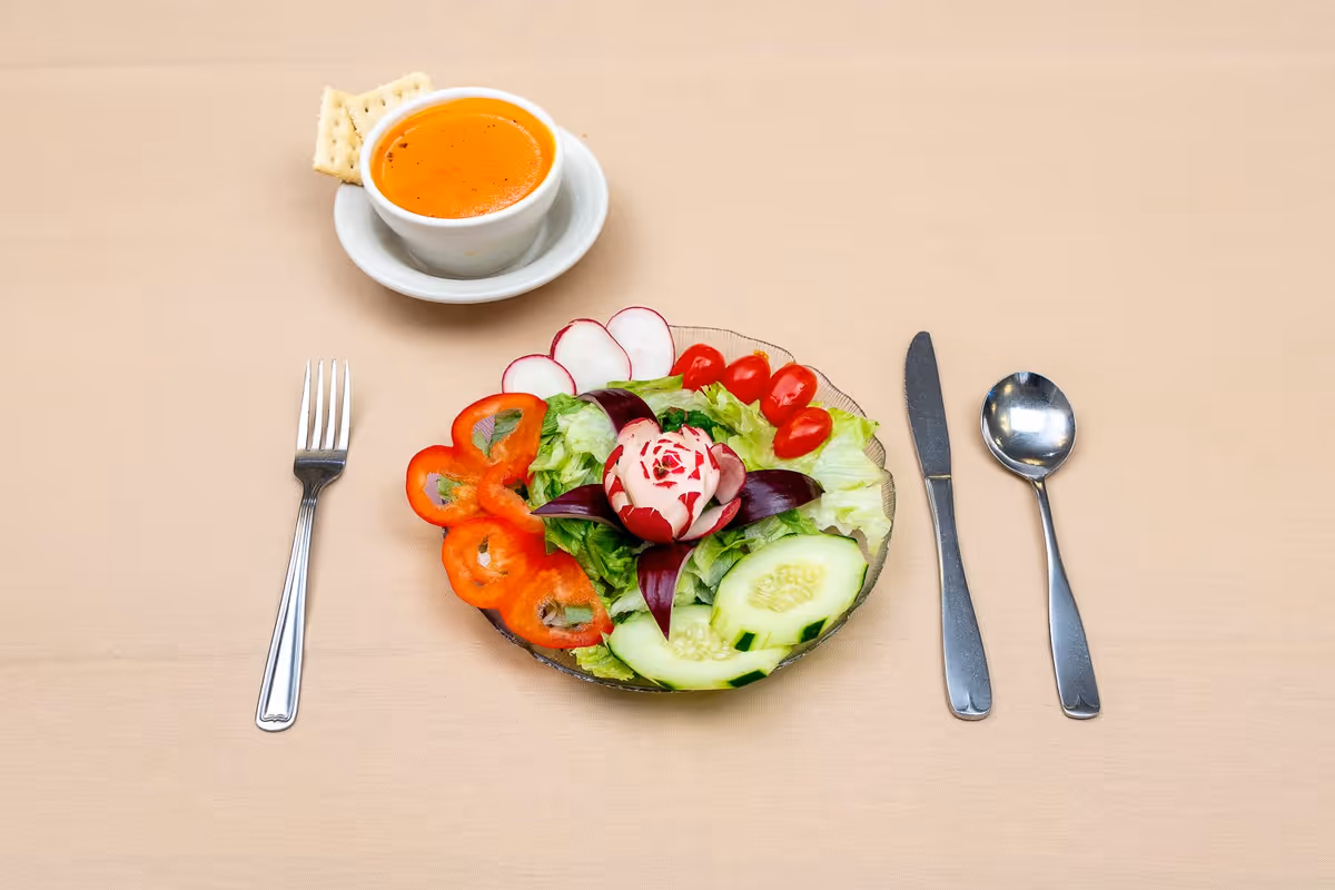 A bowl of tomato soup with two crackers on a white saucer, placed next to a plate of fresh salad consisting of lettuce, sliced cucumbers, cherry tomatoes, radishes, and red bell pepper slices, arranged on a beige tablecloth with a fork on the left and a knife and spoon on the right.