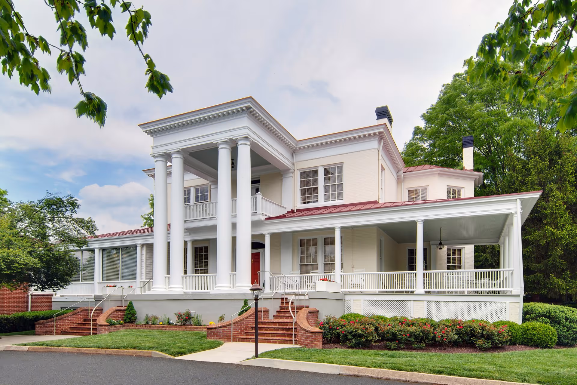 Exterior view of a large, elegant white two-story building with tall white columns, a red door, and a wrap-around porch surrounded by green trees and manicured bushes under a partly cloudy sky.