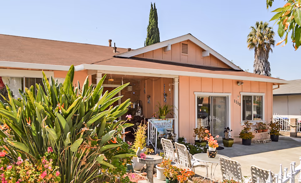 Front exterior of a pink single-story senior care home with patio seating, potted plants, and a small sign.