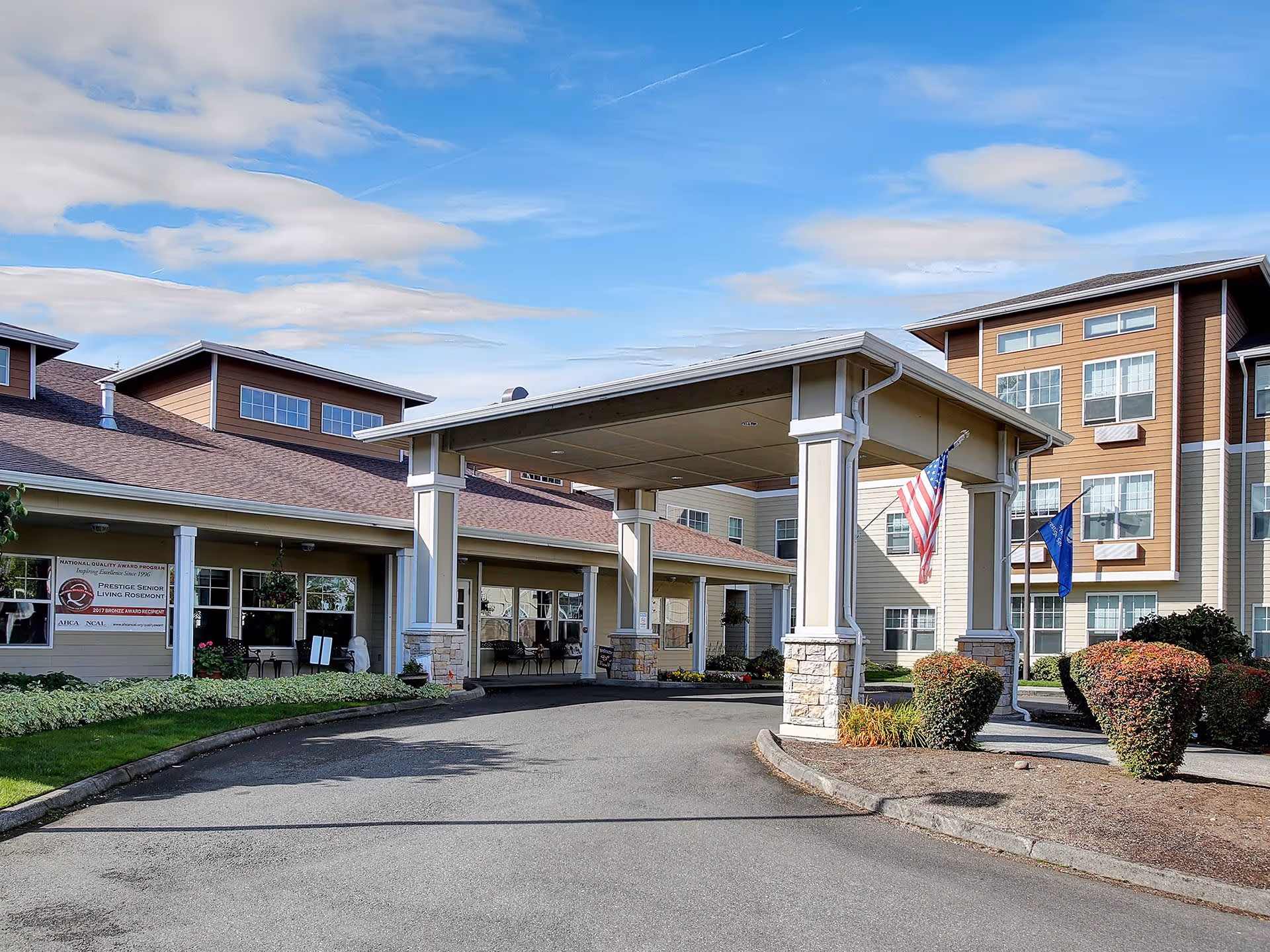 Exterior view of Prestige Senior Living Rosemont building with a covered entrance driveway, American and state flags, landscaped bushes, and a clear blue sky with some clouds.