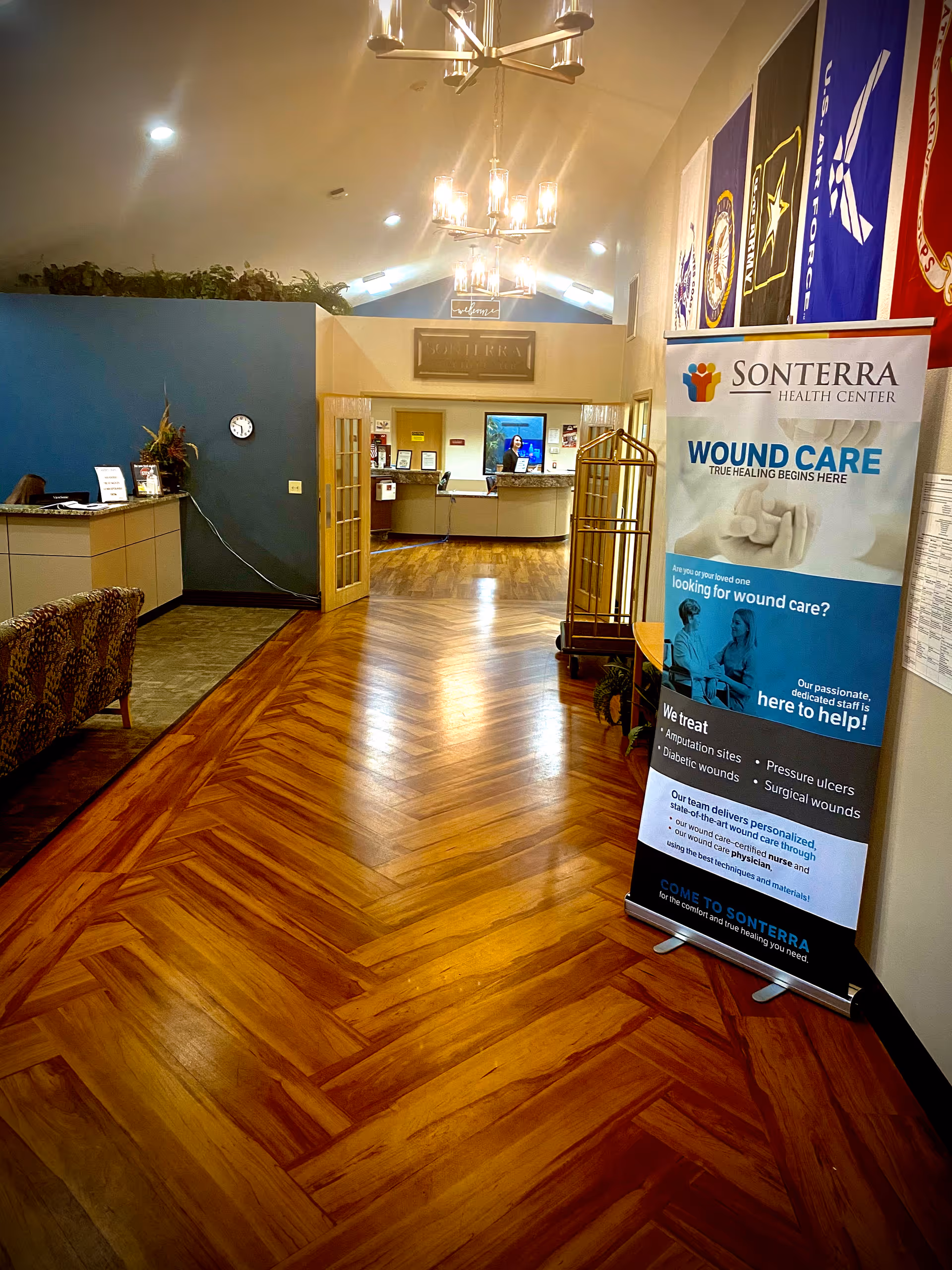 Interior view of Sonterra Health Center reception area with polished wooden floors, a seating area with patterned chairs on the left, a reception desk in the background, and a standing banner on the right promoting wound care services. Several military flags hang on the wall above the banner.
