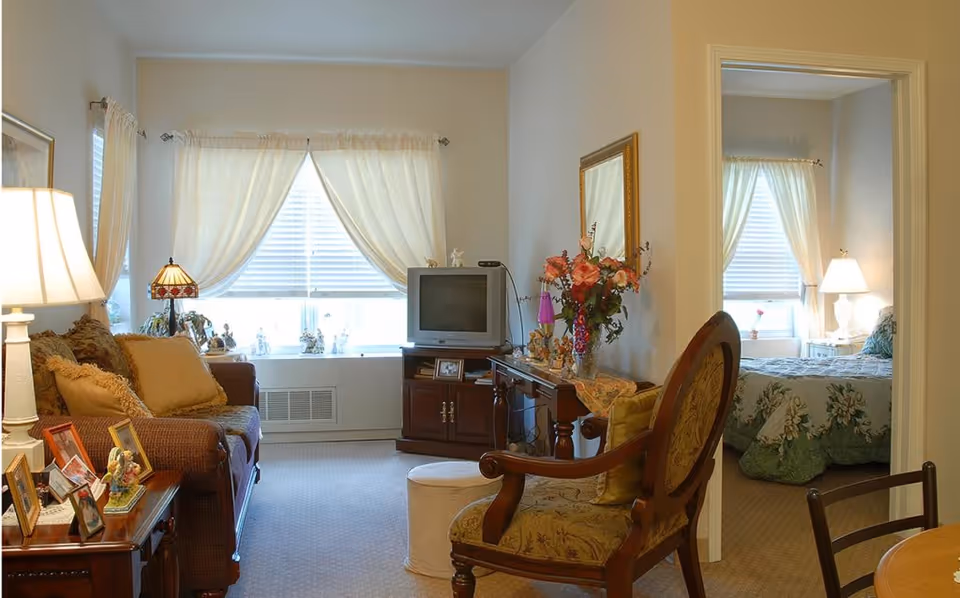 Sunny living room with a sofa, armchair, television on a cabinet, side tables with lamps and flowers, and a bedroom visible through an open doorway.