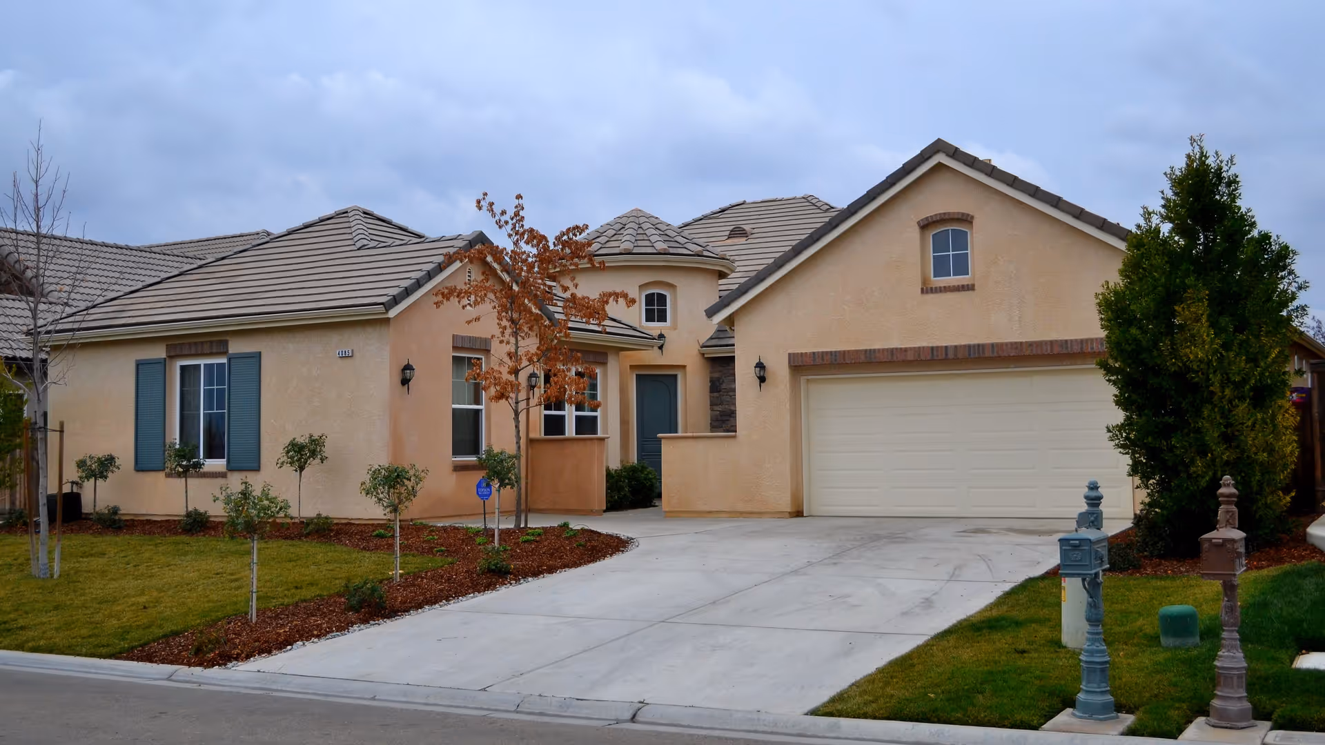 Exterior view of a single-story residential home with beige stucco walls, a tiled roof, a two-car garage, a concrete driveway, and a small landscaped front yard with young trees and shrubs under a cloudy sky.