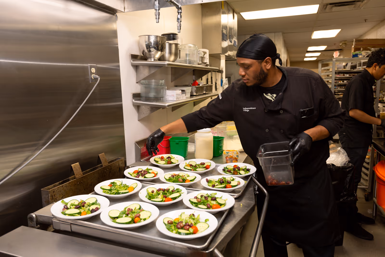 A chef wearing a black uniform and gloves is preparing multiple plates of salad in a commercial kitchen. The salads contain lettuce, cucumber slices, cherry tomatoes, and olives. Another person is working in the background in the kitchen.