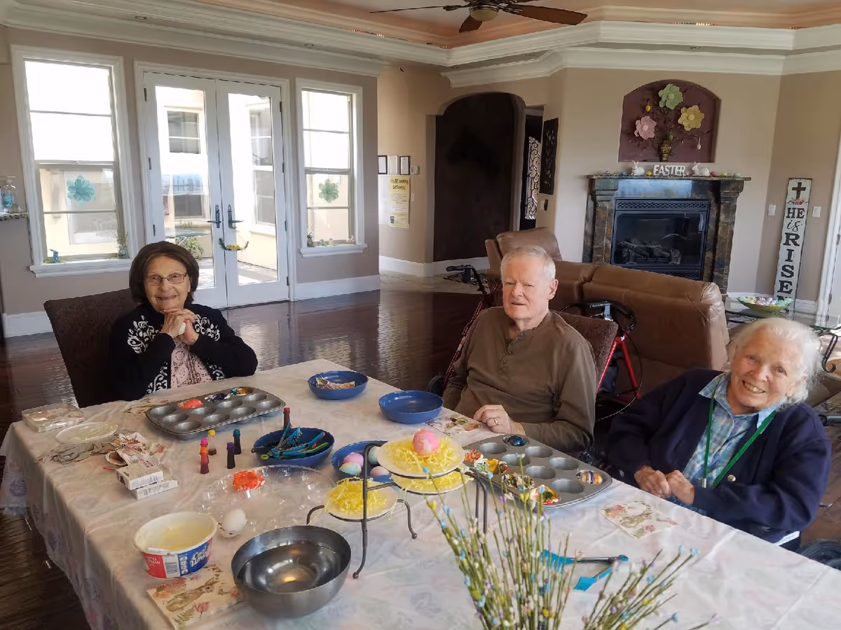 Three elderly individuals sitting around a table in a bright living room, engaging in an Easter-themed craft activity with colored eggs and decorating supplies. The room features large windows, a fireplace with Easter decorations, and comfortable seating.